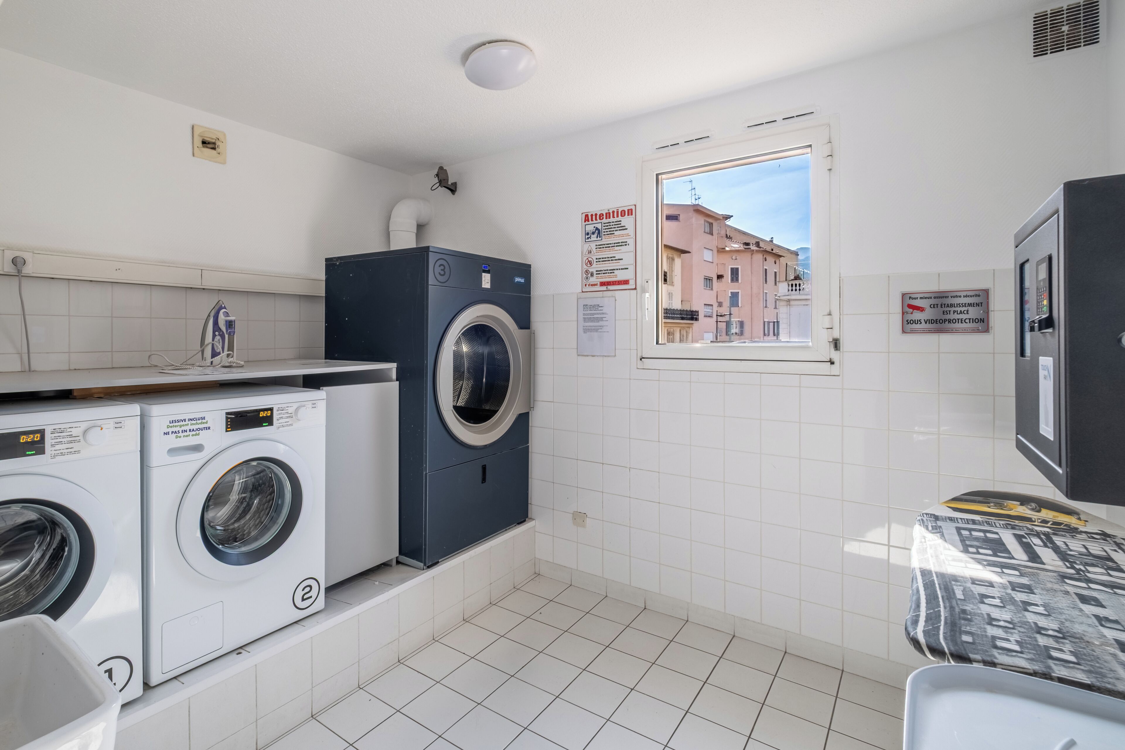 A laundry room with two washing machines, a drying machine, a window with a view of buildings, and a sign on the wall.