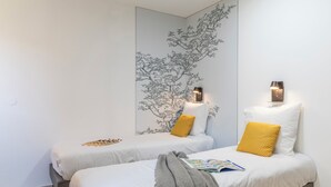 Ein Zimmer mit zwei Betten, jeweils mit weißer Bettdecke und einem Kopfkissen. Über jedem Bett befindet sich eine Wandleuchte.