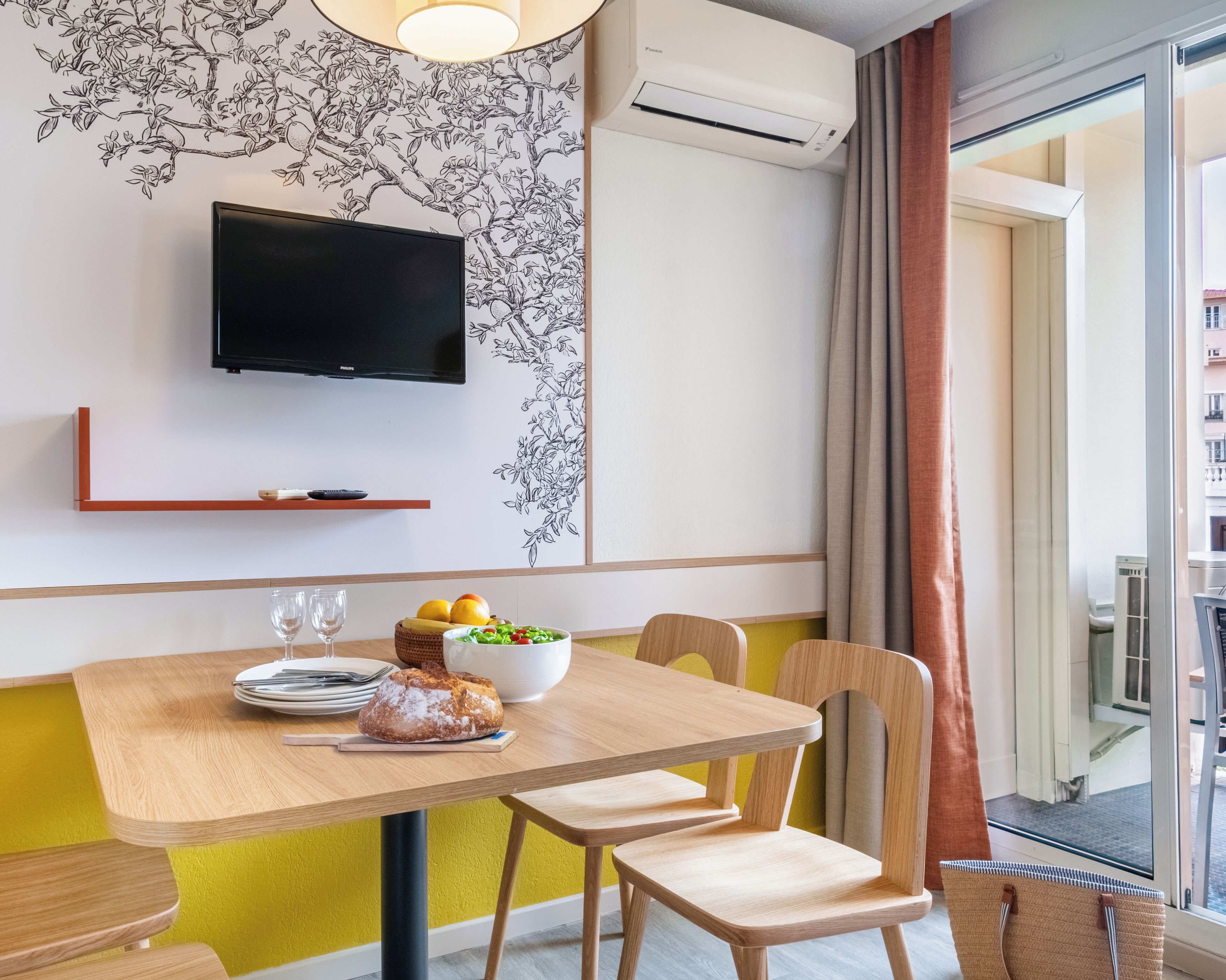 A dining area with a table set for two, a flat-screen TV mounted on the wall, and a window with curtains.