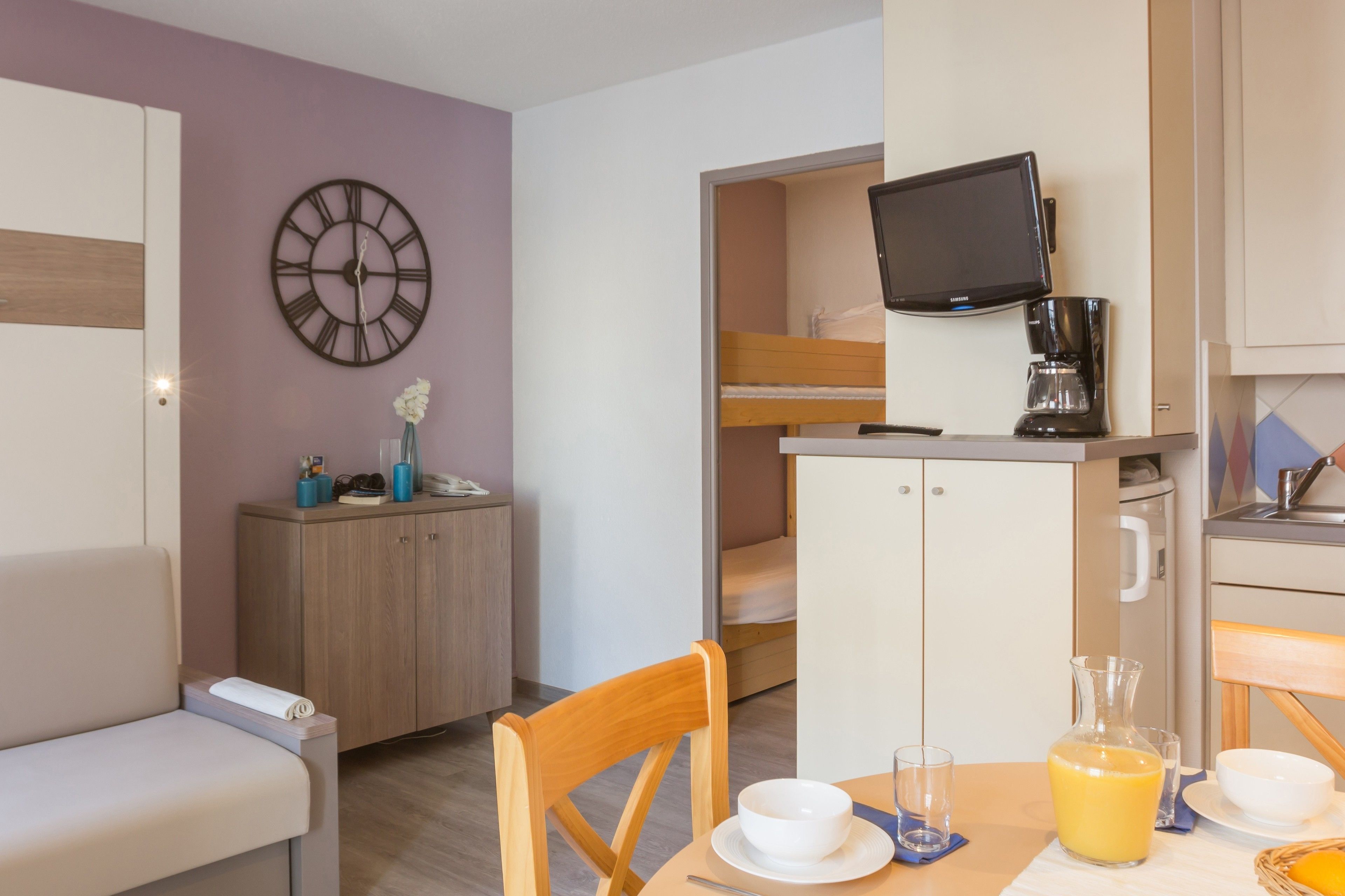 Une petite chambre comprenant un lit superposé, une télévision, une machine à café, une table à manger avec une carafe de jus d’orange et une horloge au mur.