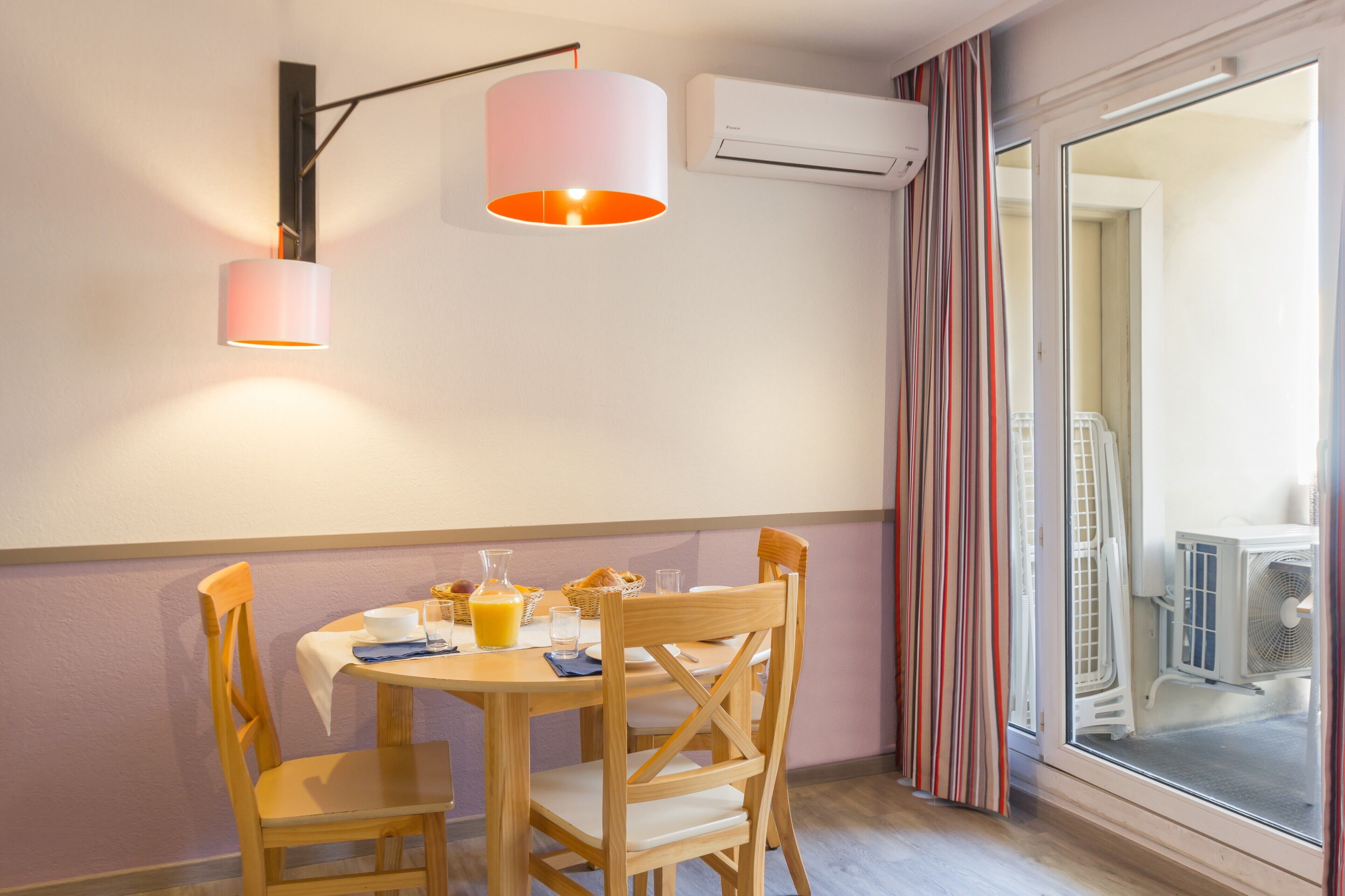 Un comedor con una mesa preparada para dos personas, un aire acondicionado de pared y una ventana con cortinas.
