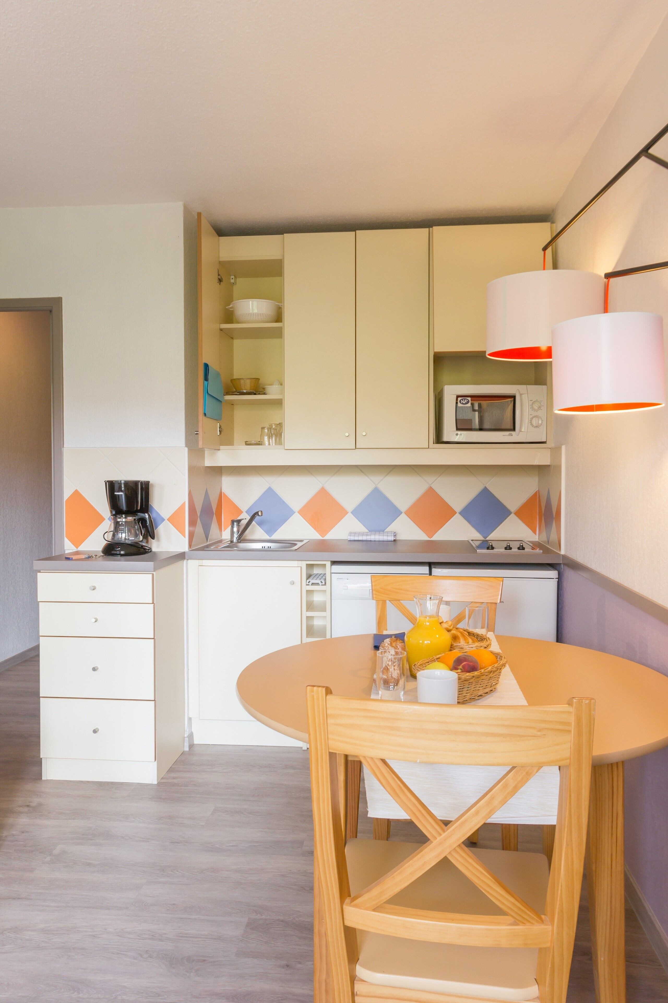 A compact kitchen with a microwave, coffee maker, and a dining table with a basket of fruit.