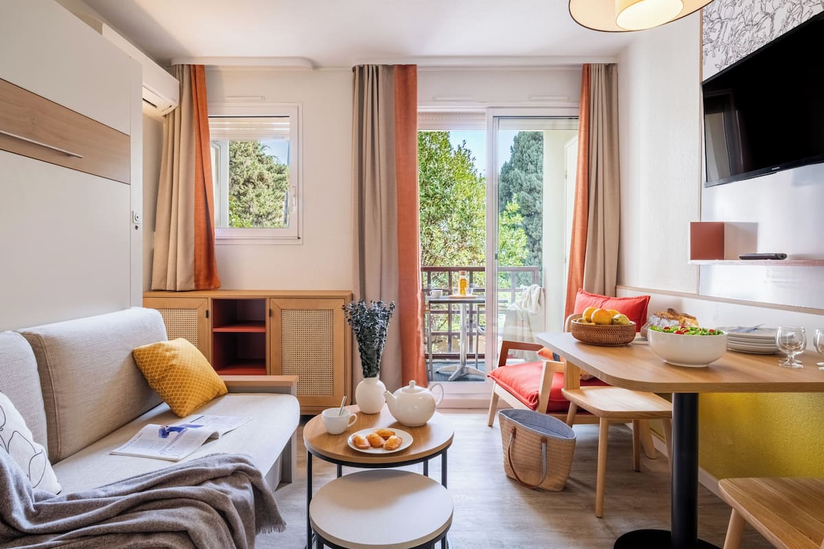 Een gezellige woonkamer met een bank, een eettafel met een schaal fruit, een bijzettafeltje met een theepot en een aan de muur bevestigde televisie.