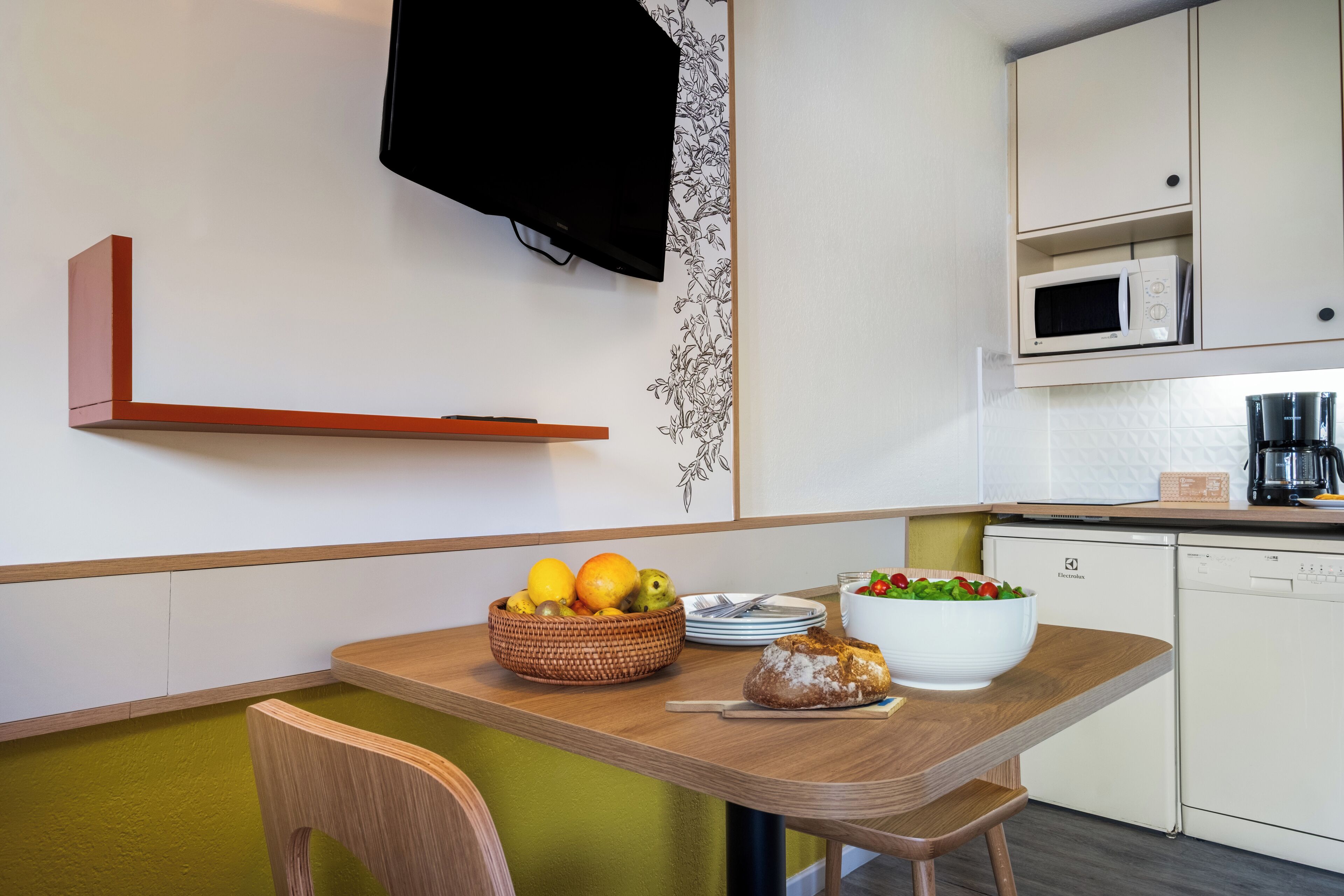 A modern kitchen with a wooden table, a fruit bowl, a white bowl with salad, and a microwave above the counter.