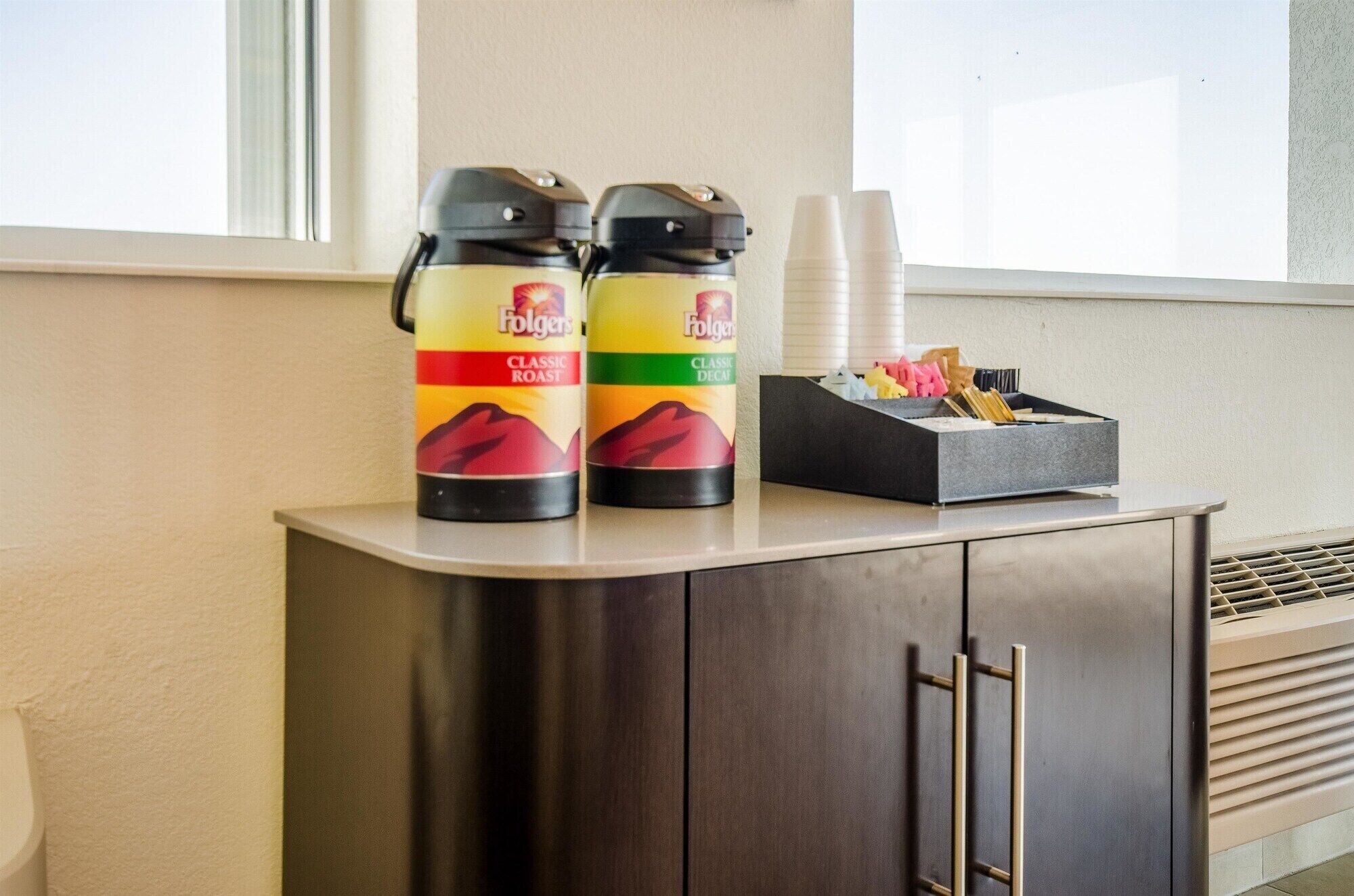 Two Folgers coffee cans on a counter, a cup holder with disposable cups, and a box with snacks.