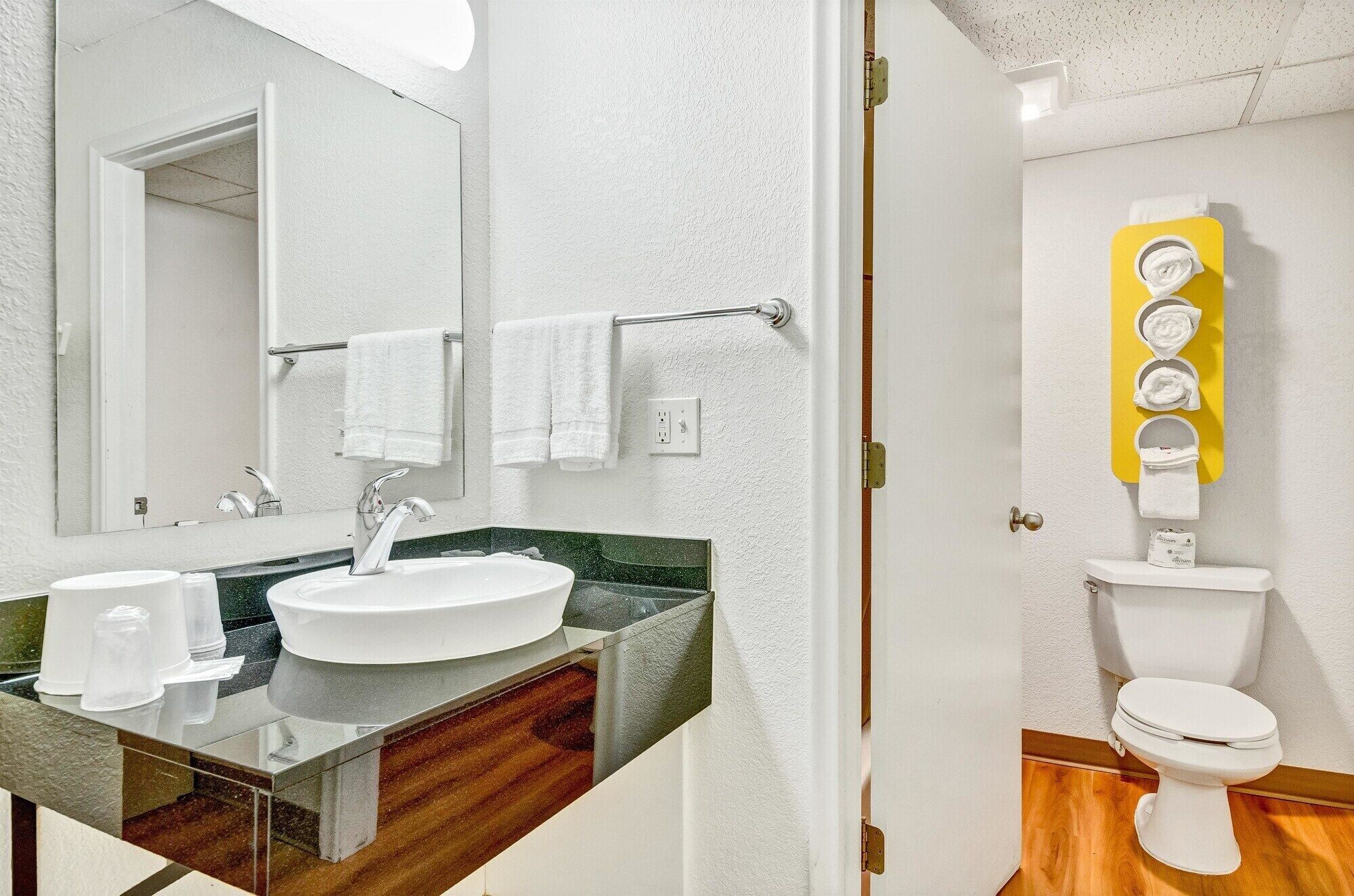 Baño con lavamanos blanco, espejo y inodoro. Hay un perchero con toallas blancas y un escurridor amarillo con varias toallas.