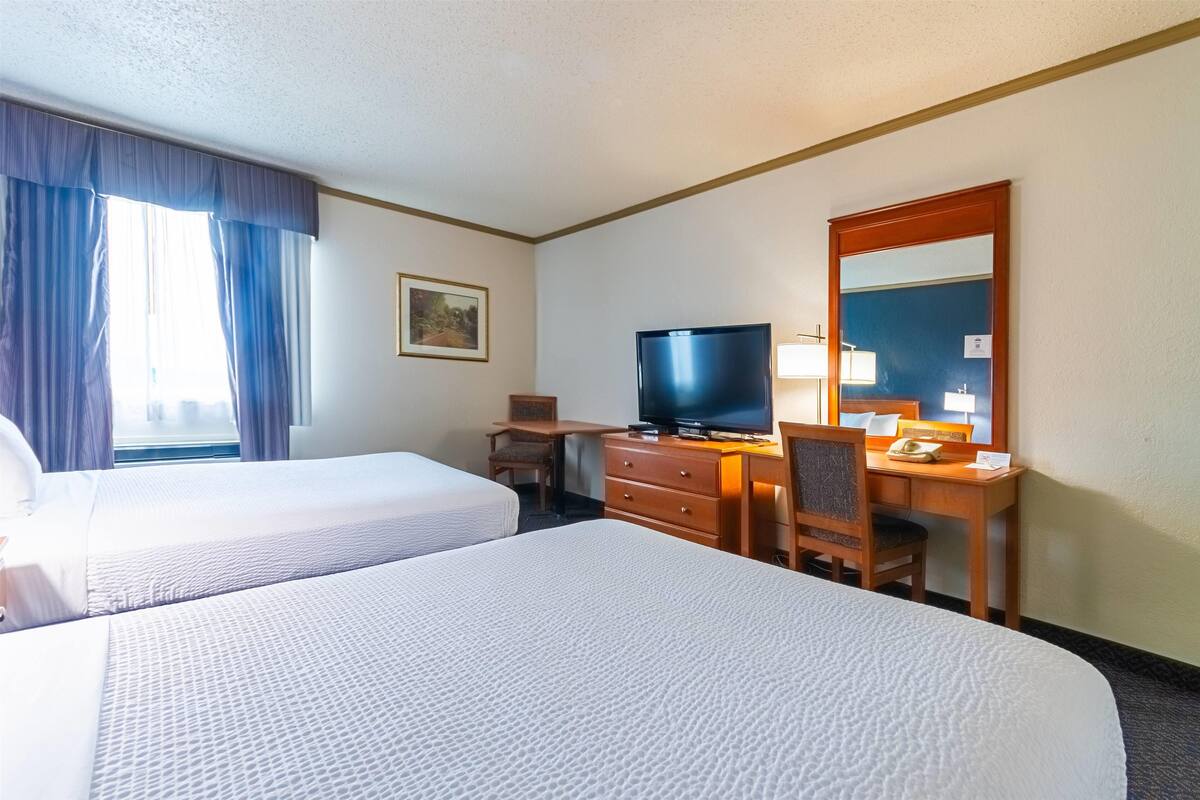 A hotel room with two beds, a desk with a television, a mirror, and a window with curtains.