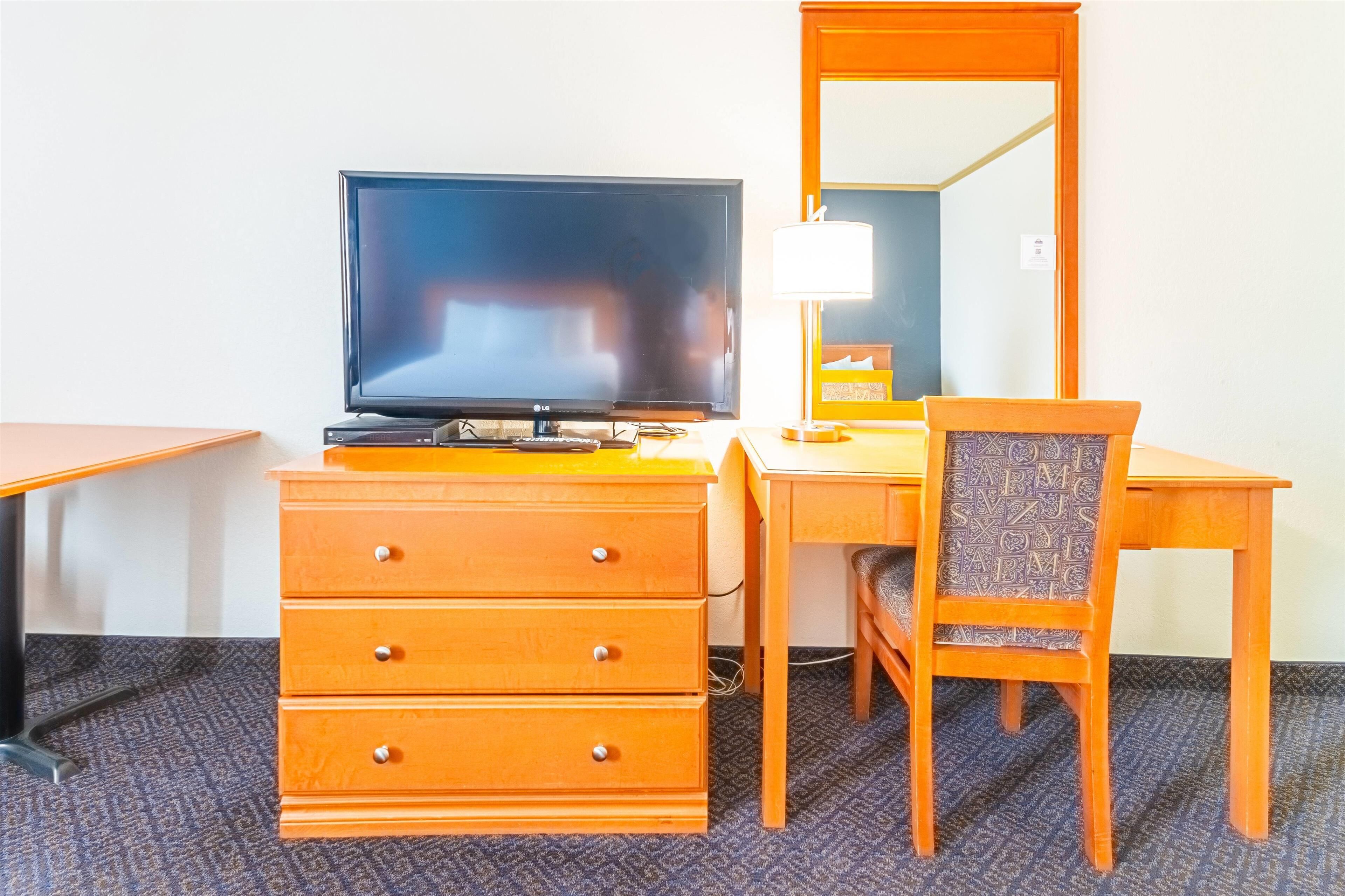 A hotel room with a wooden desk, a television, a mirror, and a chair.