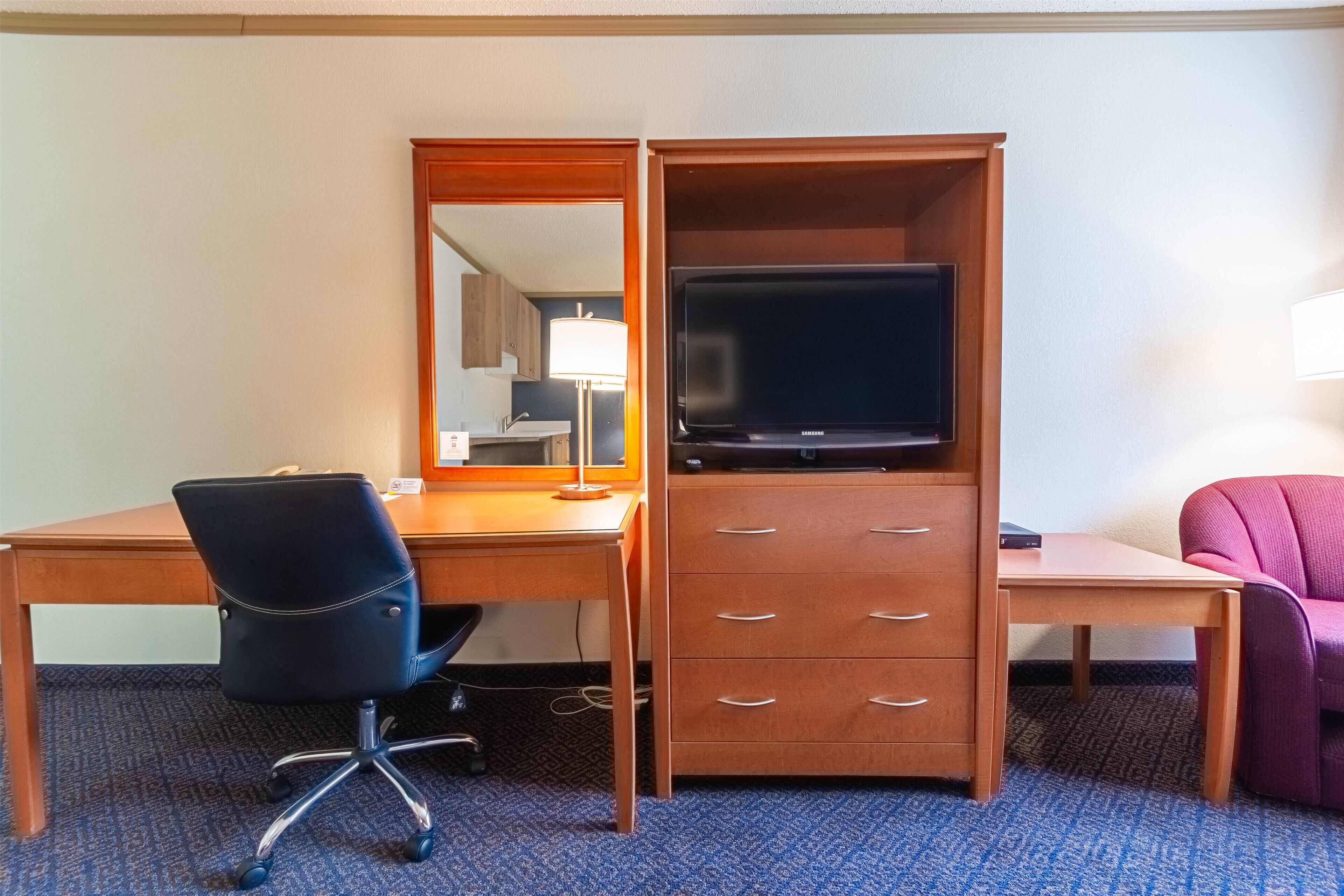 A hotel room with a wooden desk, a black office chair, a television, a wooden dresser, and a pink chair.
