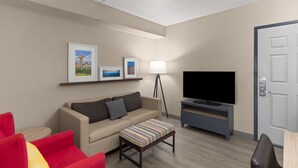 A modern living room with a sofa, a red armchair, a striped ottoman, a flat-screen TV, a floor lamp, and framed pictures on the wall.