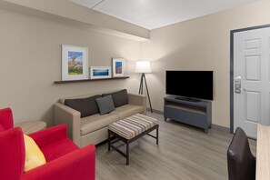 A modern living room with a sofa, a red armchair, a striped ottoman, a flat-screen TV, a floor lamp, and framed pictures on the wall. - La Quinta Inn & Suites by Wyndham Knoxville West (Knoxville)