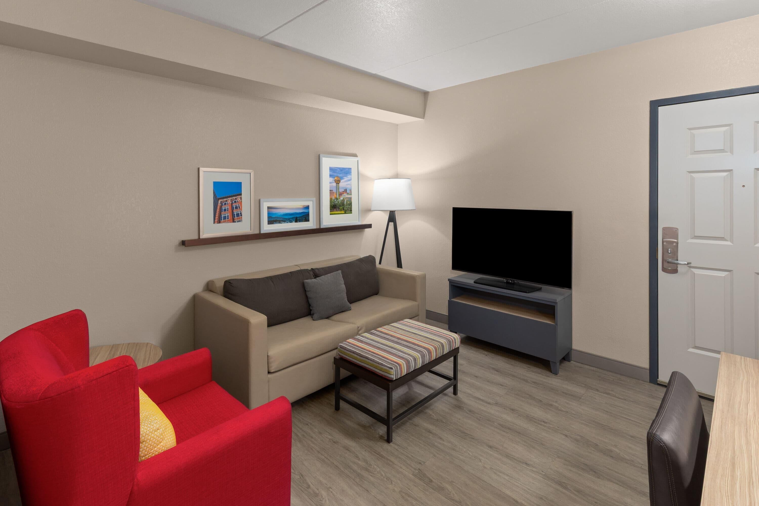 A hotel room with a sofa, a red armchair, a television, and a door.
