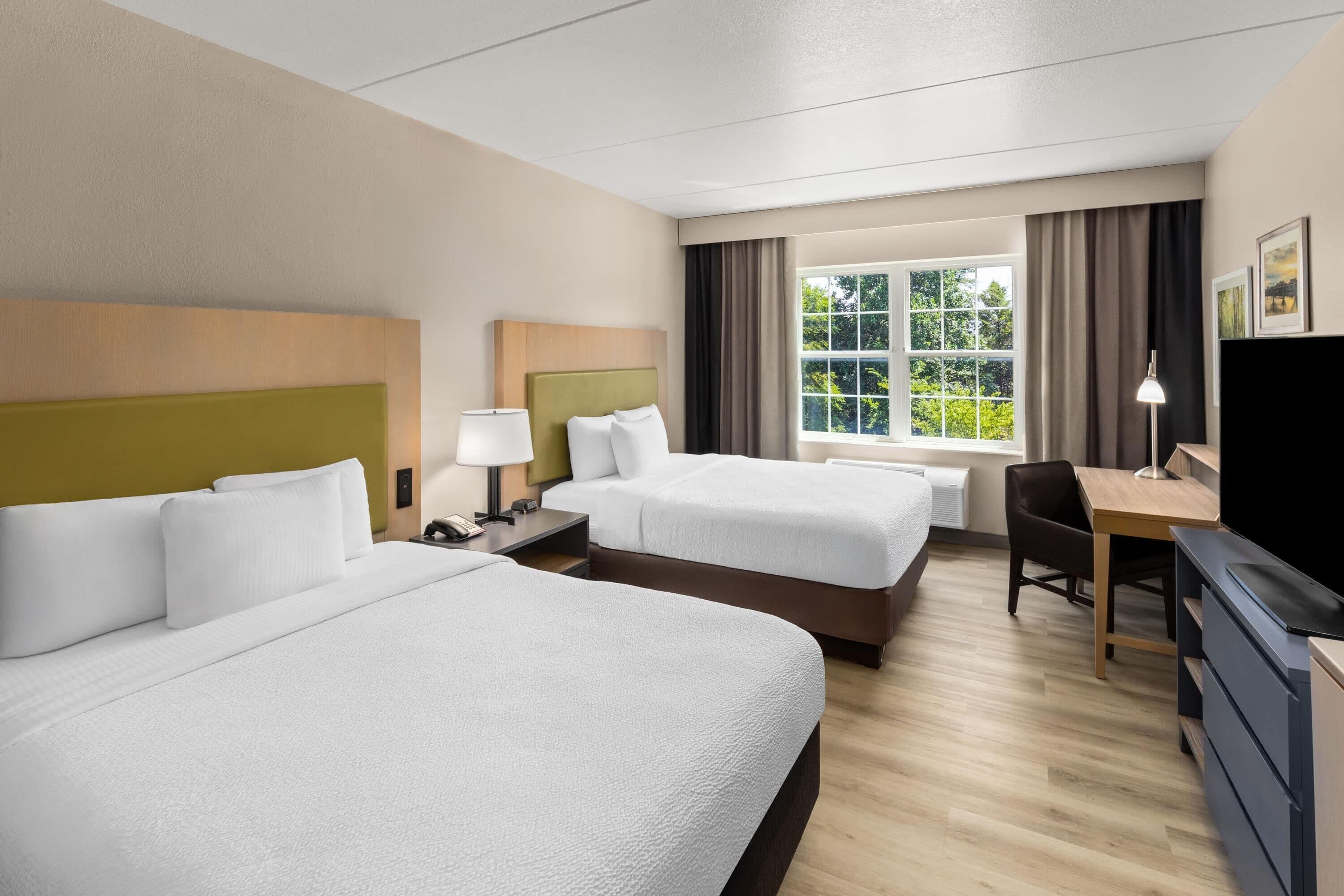 A hotel room with two beds, a desk, a chair, a TV, and a window with a view of greenery.