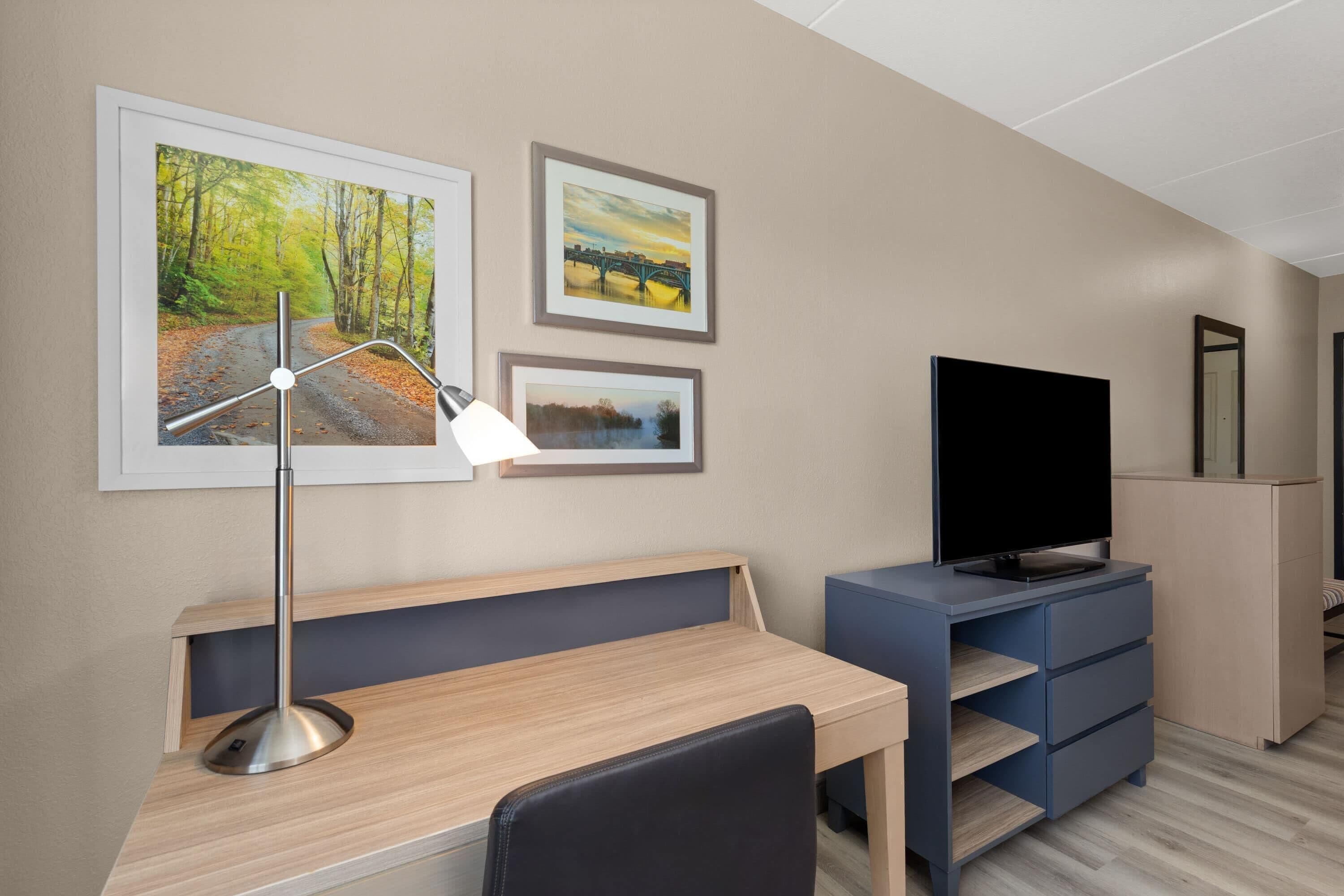 A modern hotel room with a desk, a TV, and three framed pictures on the wall.