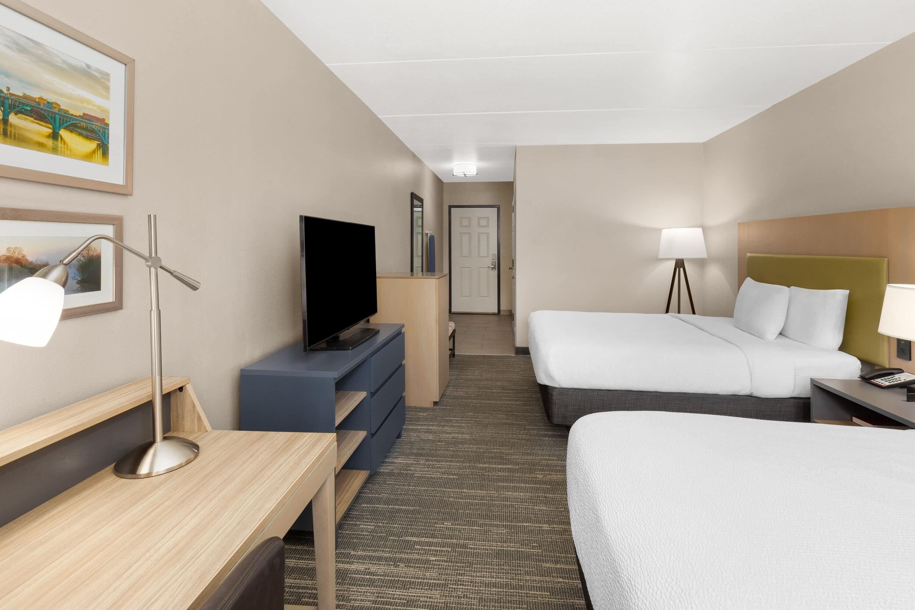A hotel room with a desk, a television, a bed, a bedside table with a lamp, and a carpeted floor.