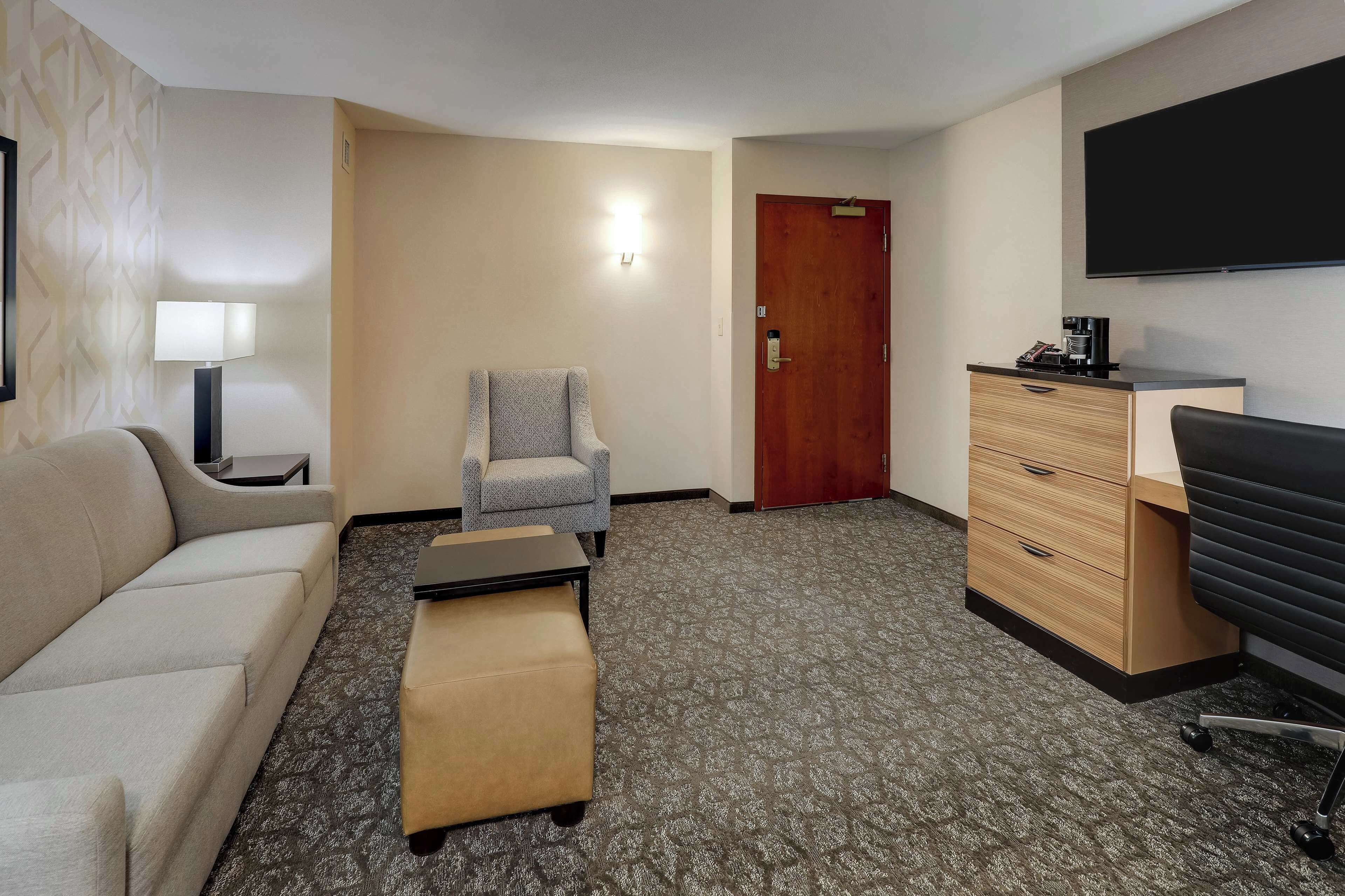 A hotel room with a sofa, armchair, ottoman, desk with a coffee maker, and a TV mounted on the wall.