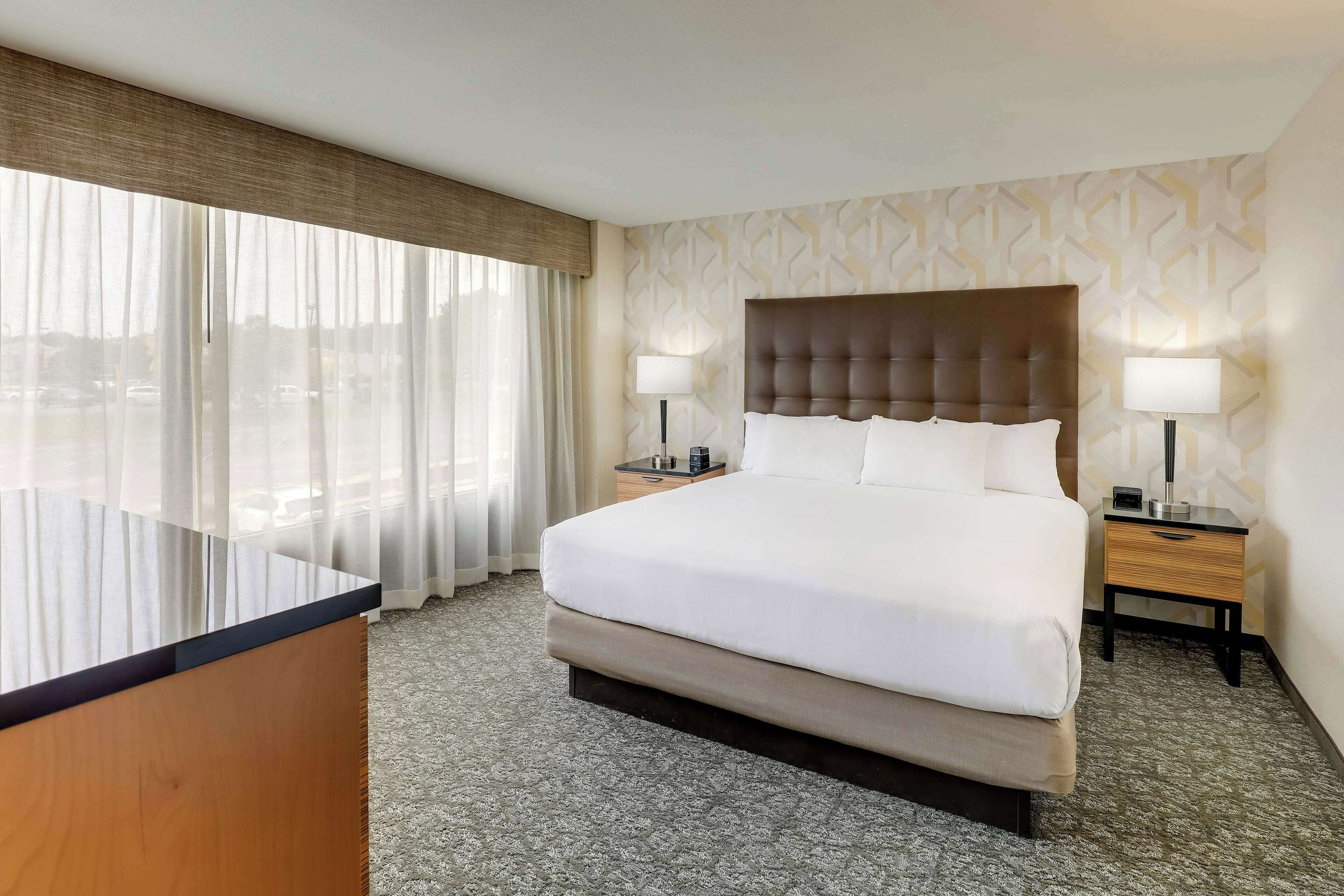 Habitación de hotel con una cama grande, dos mesitas de noche con lámparas, un escritorio y vistas de la ciudad a través de cortinas translúcidas.