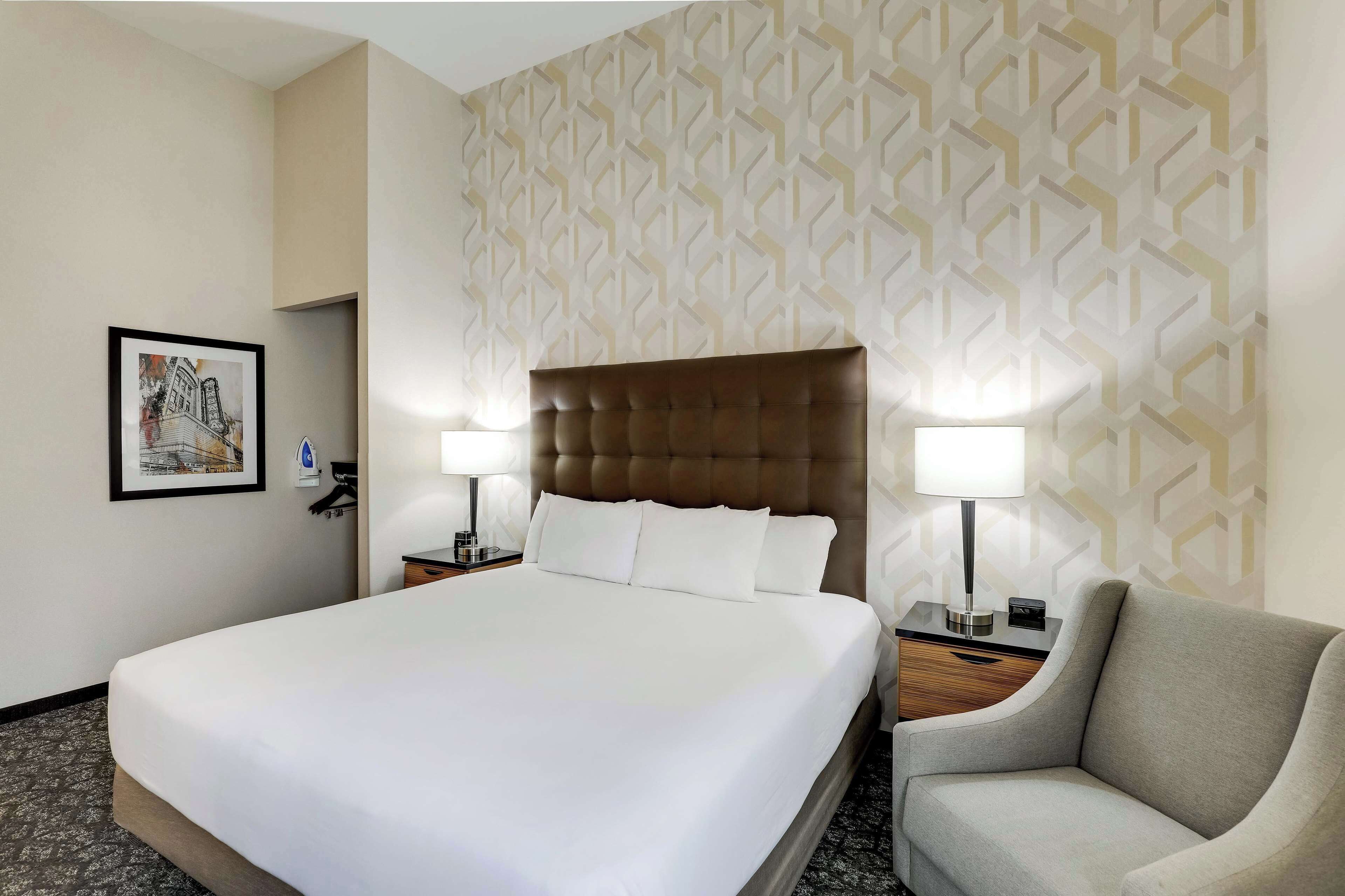 A hotel room with a large bed, two bedside lamps, a chair, and a framed picture on the wall.