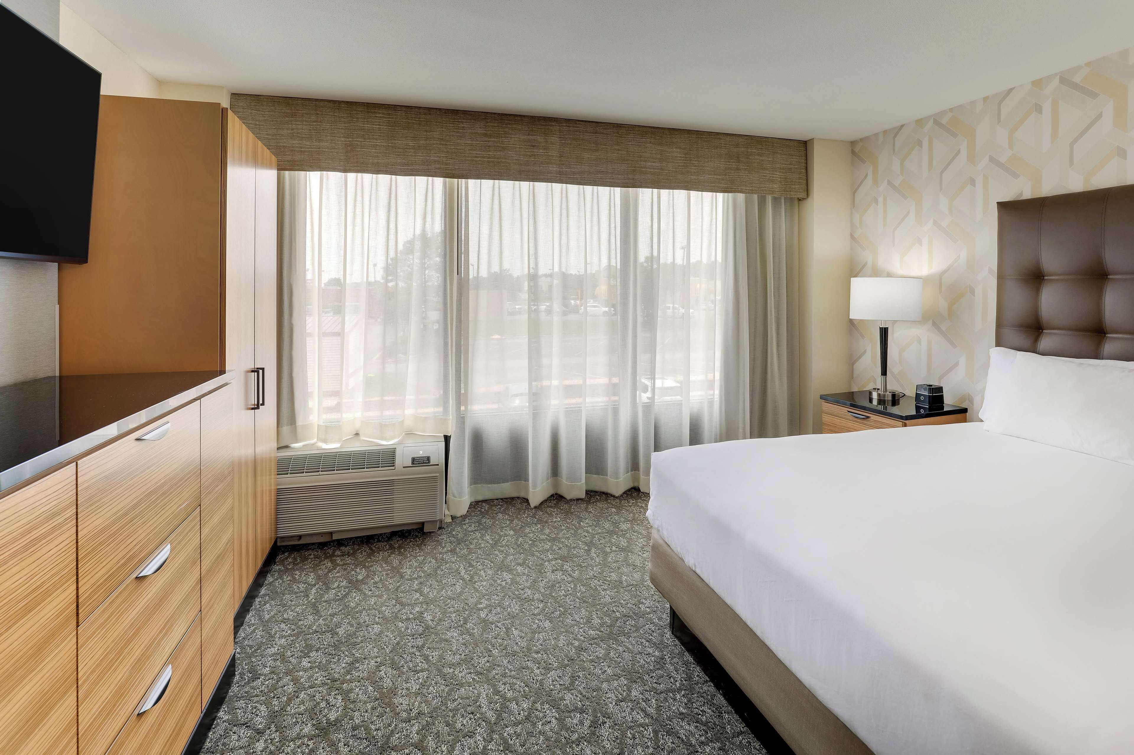 A hotel room with a large bed, a flat-screen TV, a desk with a lamp, and a view of the city through sheer curtains.