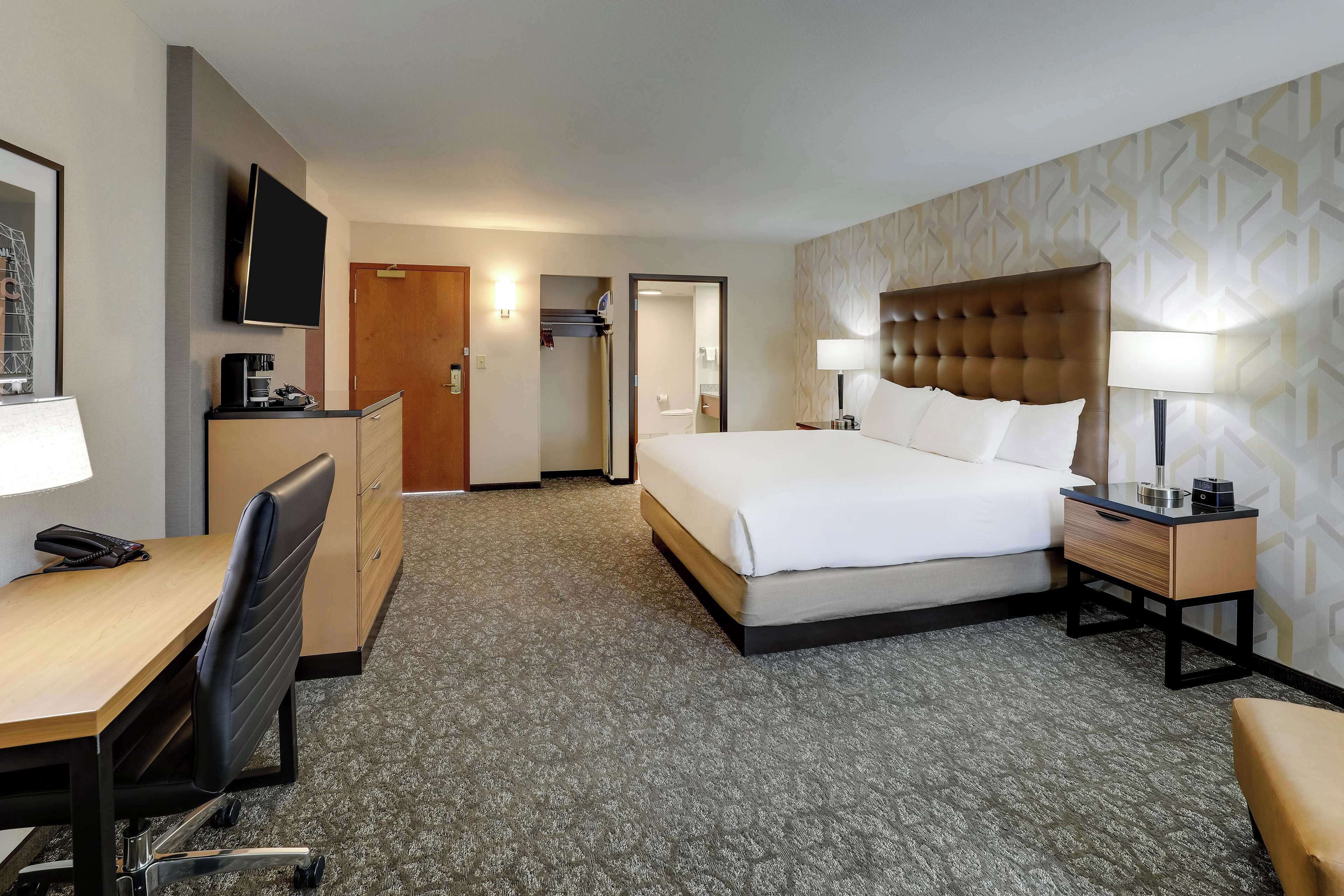 Une chambre d’hôtel avec un lit, un bureau, une chaise, une télévision et une salle de bain visible à travers une porte ouverte.