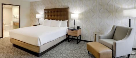 A hotel room with a large bed, a bedside table with a lamp, a chair, and a small ottoman.