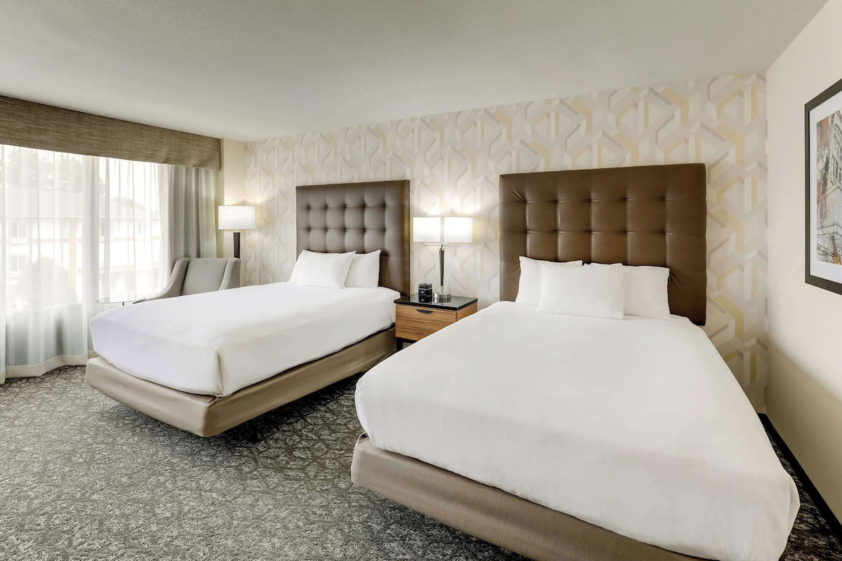 Habitación de hotel con dos camas, un ventanal grande con cortinas de gasa, un papel tapiz estampado y un cuadro en la pared.