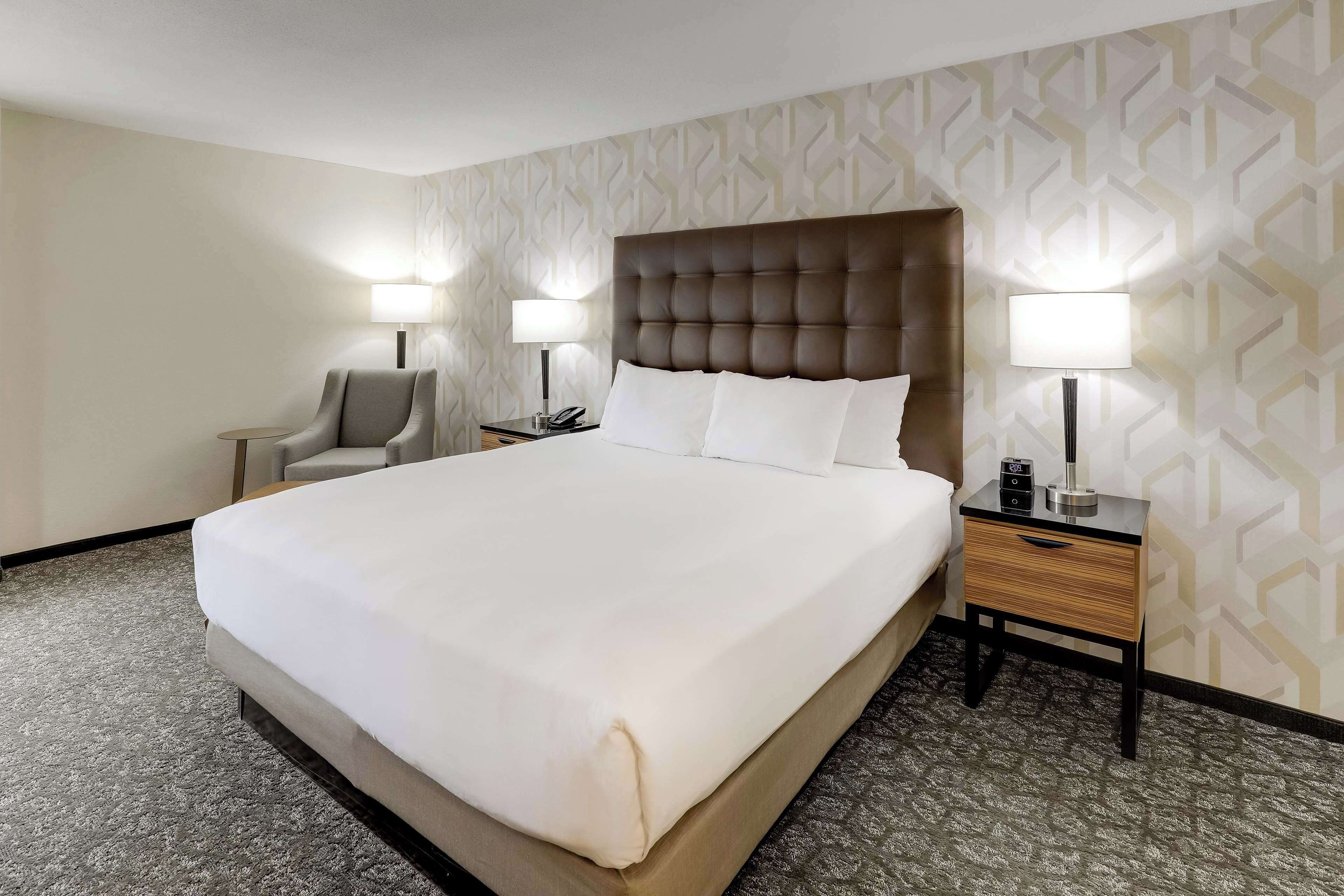 A hotel room with a large bed, two bedside tables with lamps, a chair, and a patterned wall.