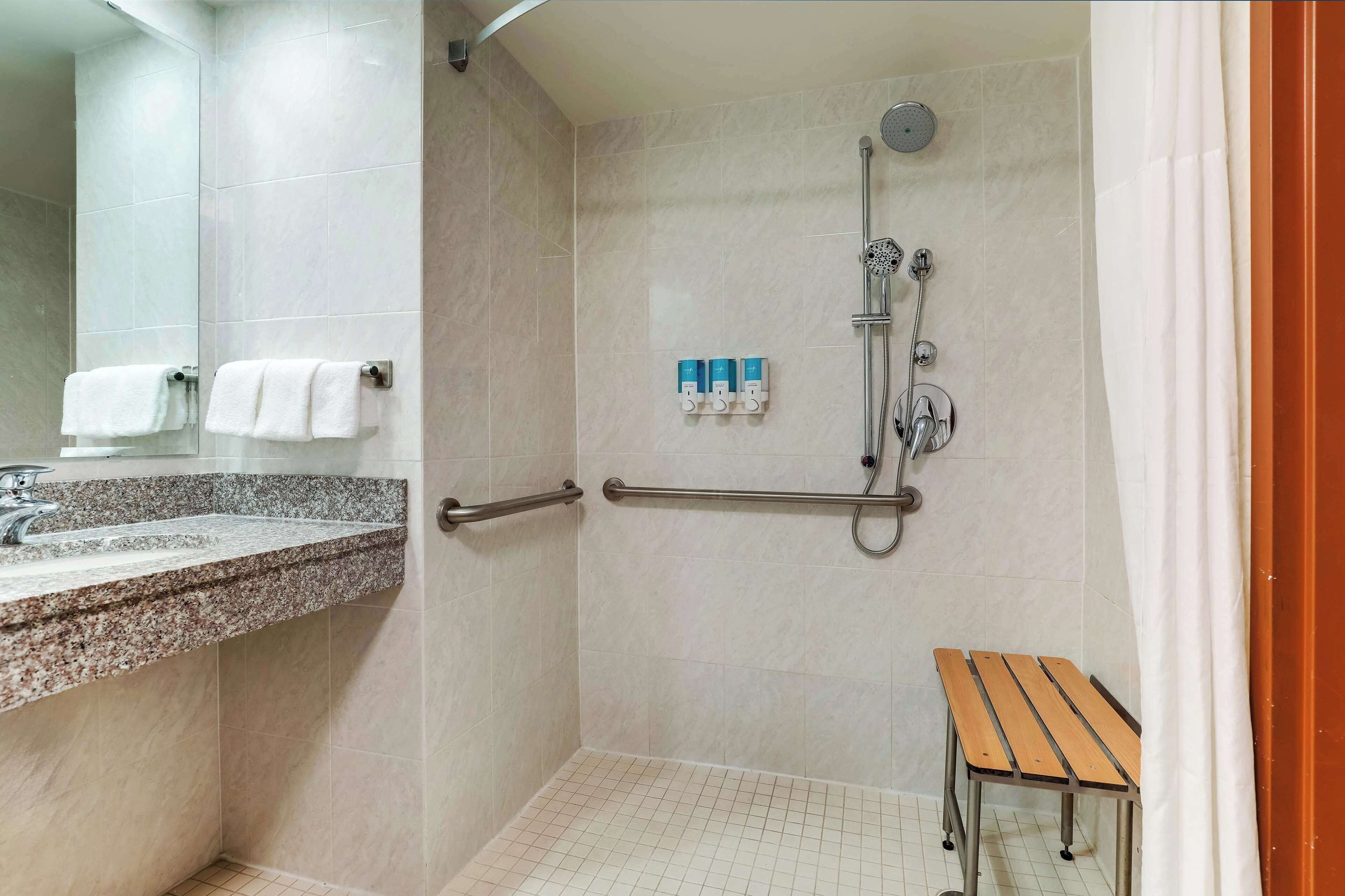 Baño con ducha, barras de apoyo, un banco y un lavabo con espejo.