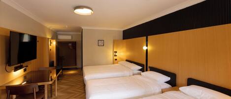 Habitación de hotel con tres camas, un escritorio con silla, una televisión y una lámpara fijada a la pared.