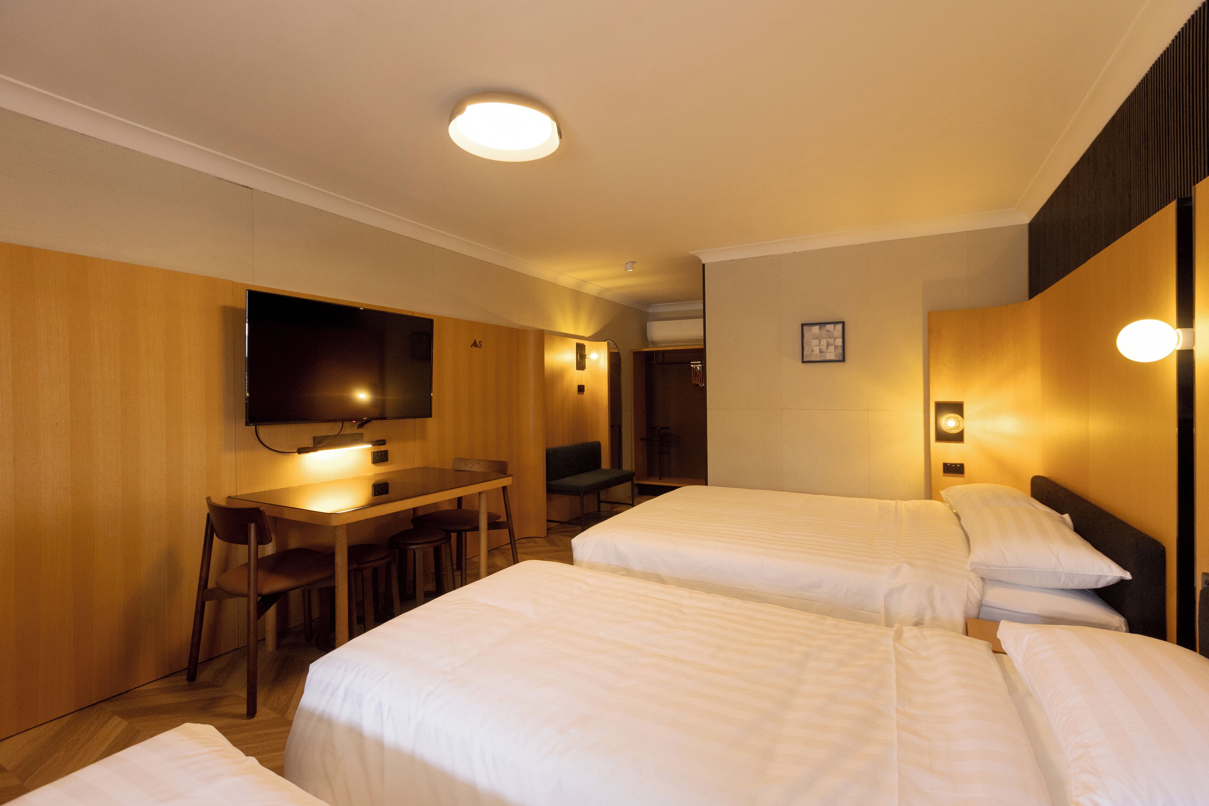 A hotel room with two beds, a desk, chairs, a TV, and a wall-mounted light fixture.