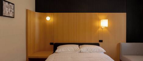 Una habitación de hotel con cama, mesita de noche, una silla y dos lámparas de pared.