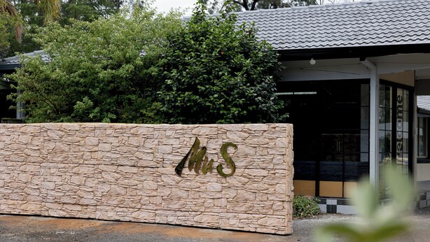 A stone wall with the inscription "MUS" in the foreground, a modern building with a tiled roof, and a tree in the background. - Mu S Boutique Motel (Coonabarabran)