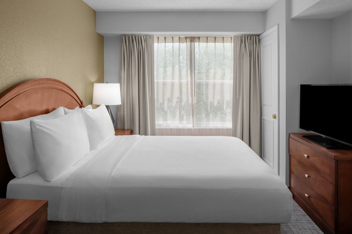 Quarto de hotel com uma cama grande, televisão sobre uma cômoda de madeira, mesa de cabeceira com abajur e janela com cortinas.