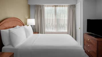 Habitación de hotel con una cama grande, un televisor en una cómoda de madera, una mesita de noche con una lámpara y una ventana con cortinas.