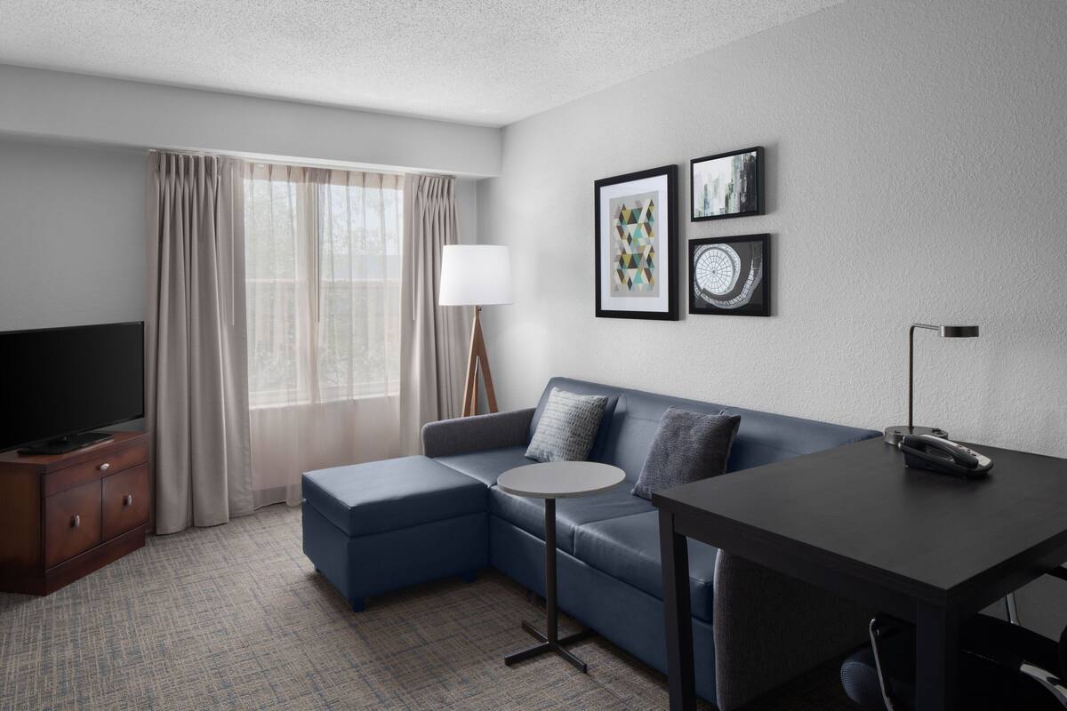 Quarto de hotel moderno com sofá modular, uma mesinha redonda, televisão em móvel de madeira e obras de arte na parede.