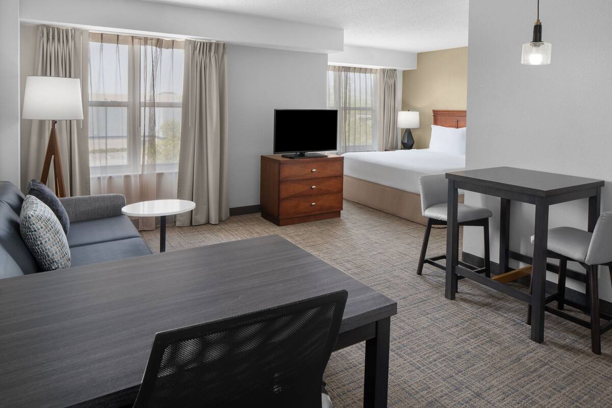 Quarto de hotel com cama, televisão em cômoda de madeira, mesa de jantar com cadeiras e um sofá.