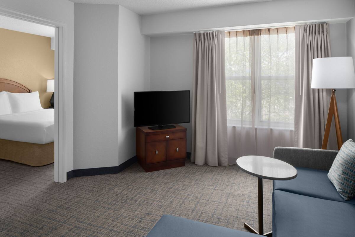 Quarto de hotel com uma cama, televisão num móvel de madeira, um sofá, uma mesinha e um abajur.