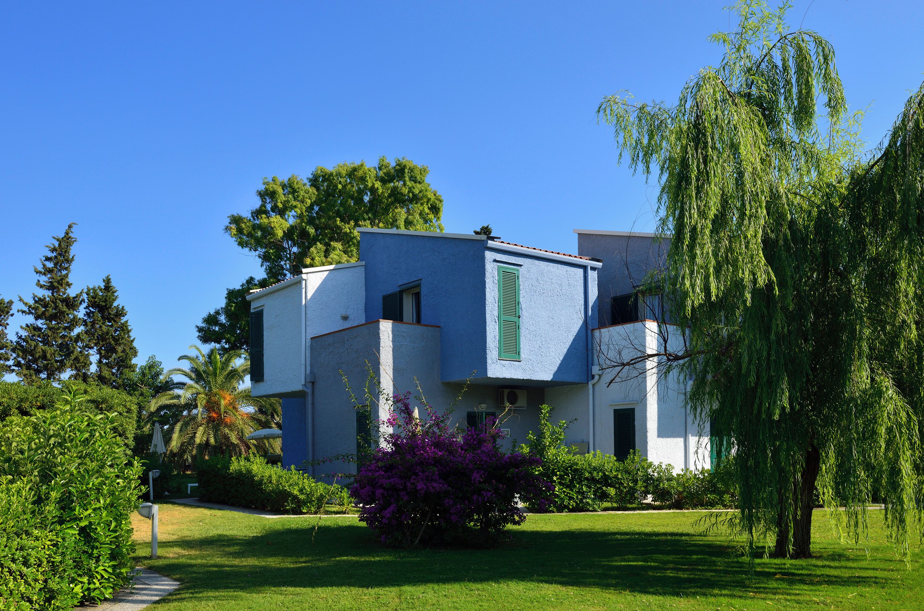 A modern two-story building with a flat roof, surrounded by lush greenery and a clear blue sky.