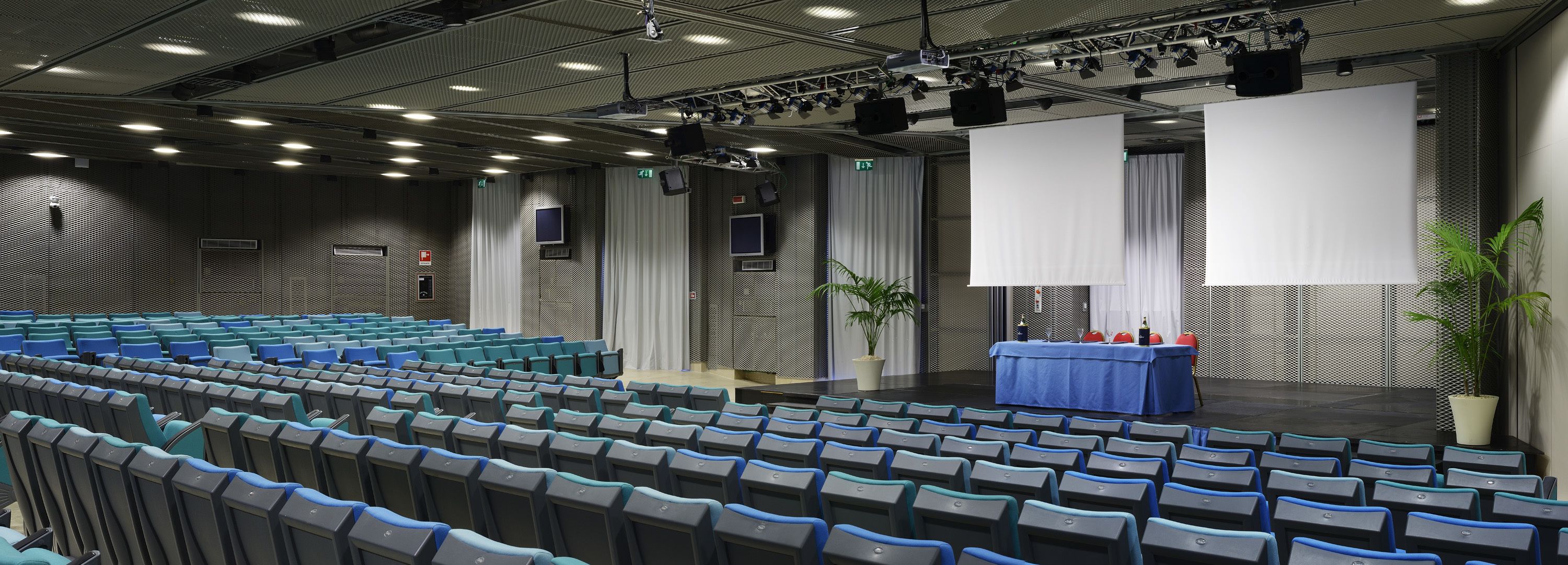An auditorium with rows of blue and gray chairs, a stage with a blue table, and a large white screen.