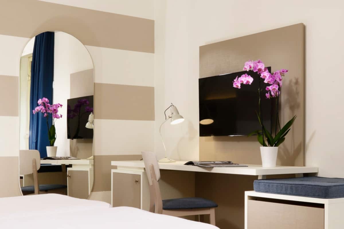 A hotel room with a bed, a desk, a chair, a desk lamp, a mirror, and a potted orchid.