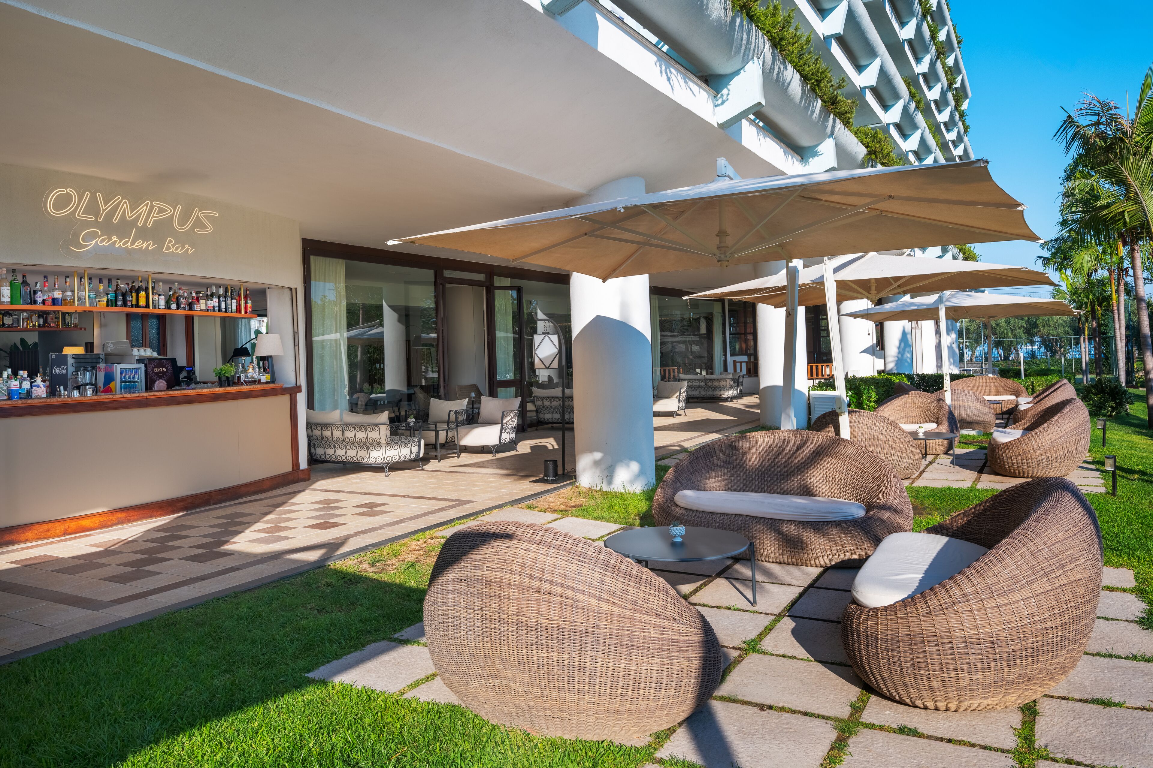 Un espace extérieur aménagé avec des meubles en osier, un bar agrémenté d’une enseigne au néon et une vue sur un bâtiment moderne.