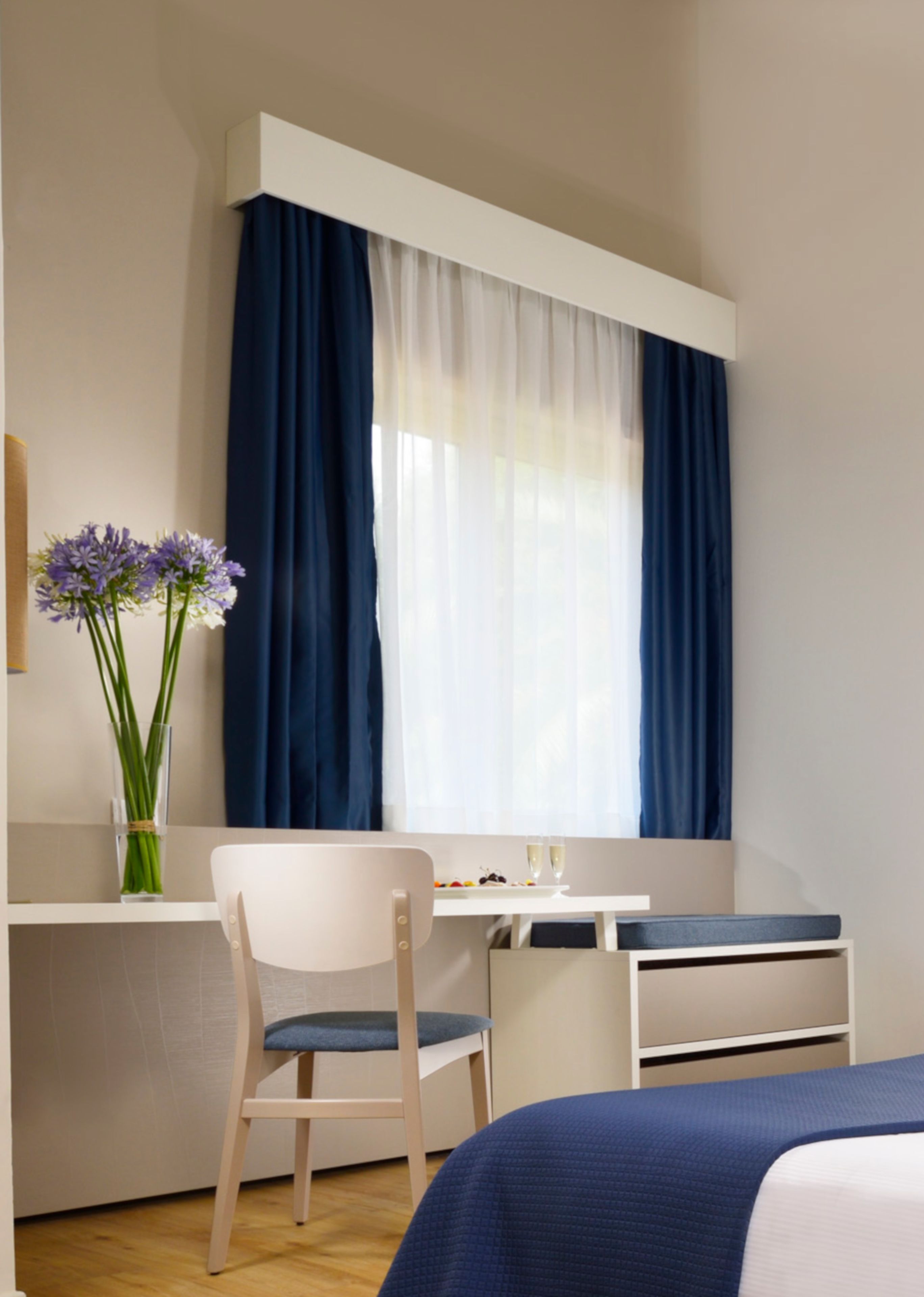 A bedroom with a bed, a desk, and a chair. There is a window with curtains, a vase with flowers, and a small table with a plate of fruit and two glasses.
