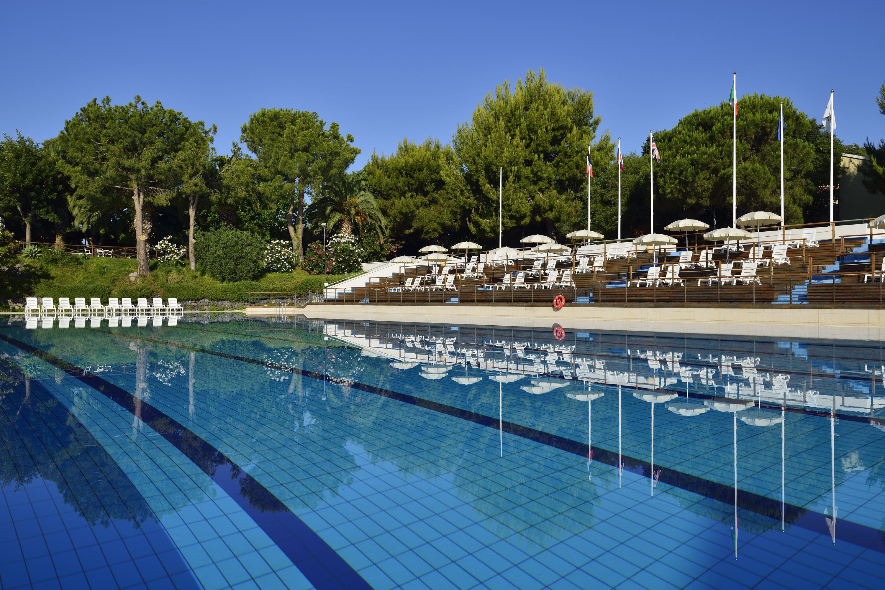 Piscina com espreguiçadeiras e guarda-sóis, rodeada por árvores e bandeiras.