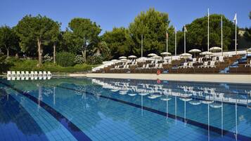 Piscina com espreguiçadeiras e guarda-sóis, rodeada por árvores e bandeiras.