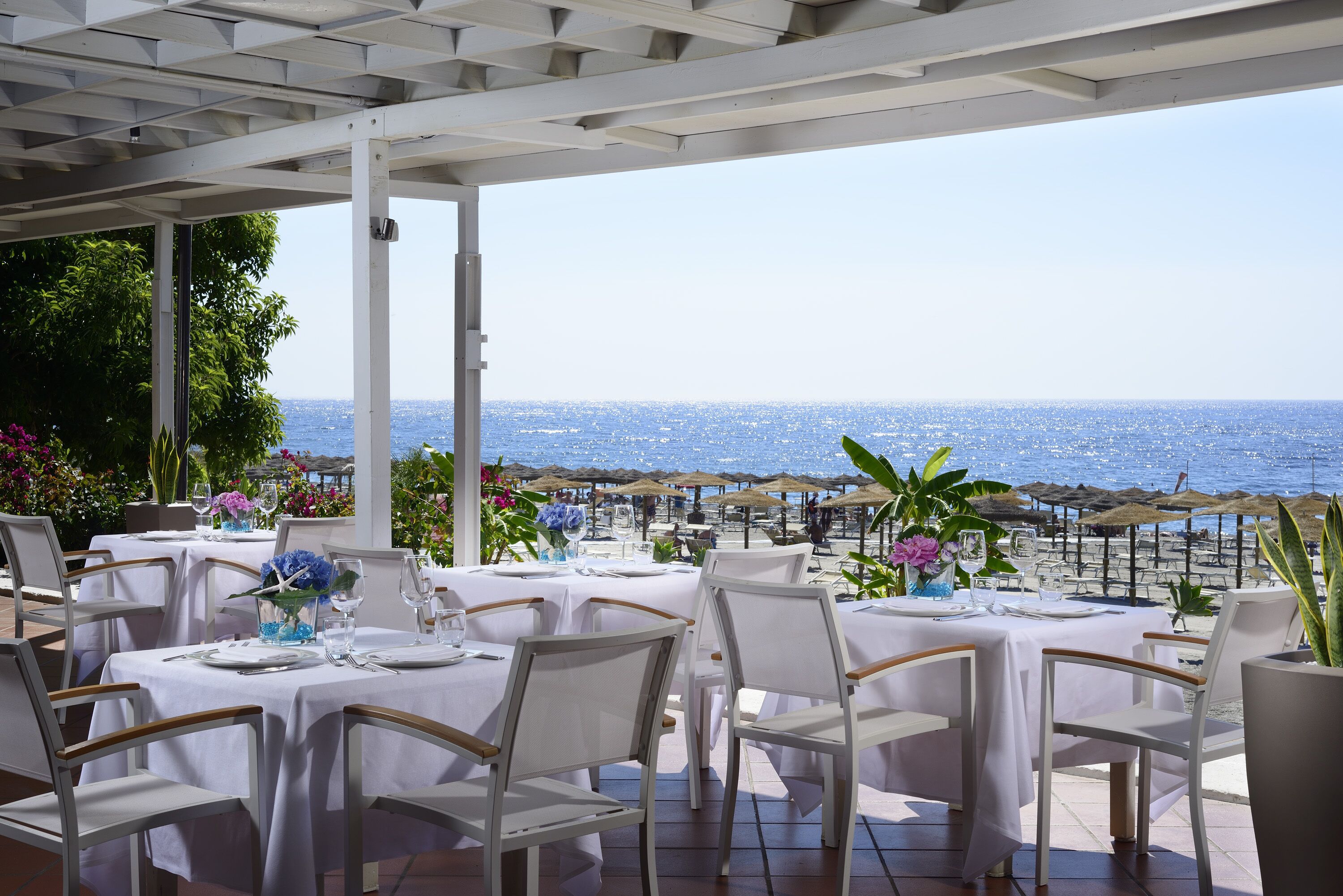 Un espace de restauration en plein air avec des tables disposées pour deux personnes, offrant une vue sur une plage et la mer.