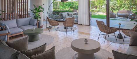 A modern hotel lobby with a sofa set, round coffee tables, wicker chairs, and a view of the outdoor garden.