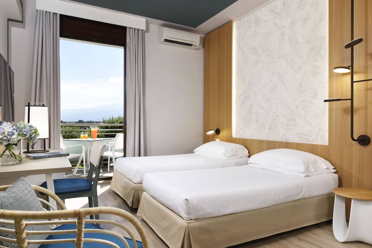 A hotel room with two beds, a desk with a chair, a small table, and a view of the outdoors.
