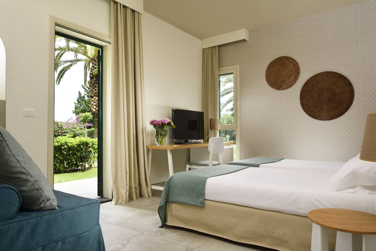 A hotel room with a bed, a desk with a TV, a chair, and a view of greenery outside.