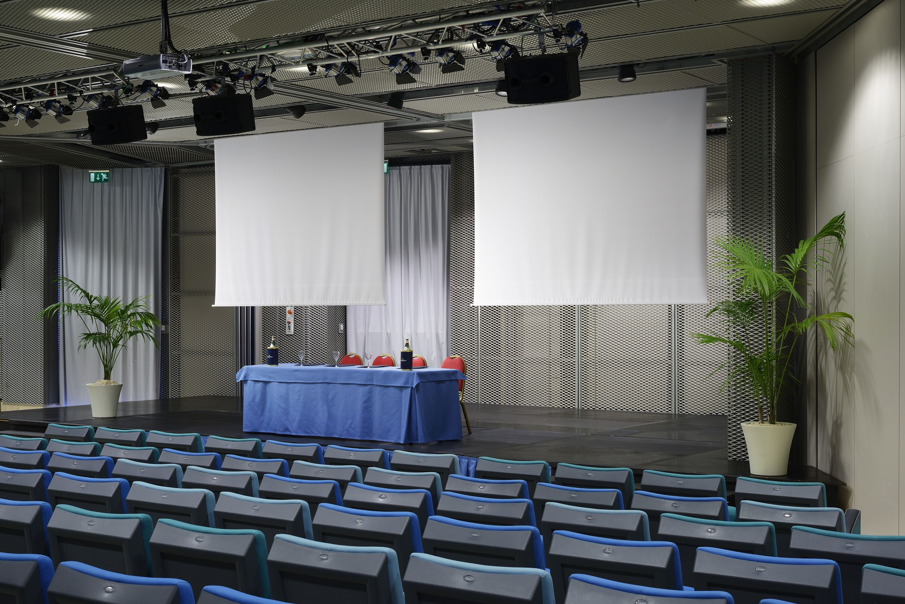 A conference room with rows of chairs, a stage with a blue table and red chairs, and two large blank screens.