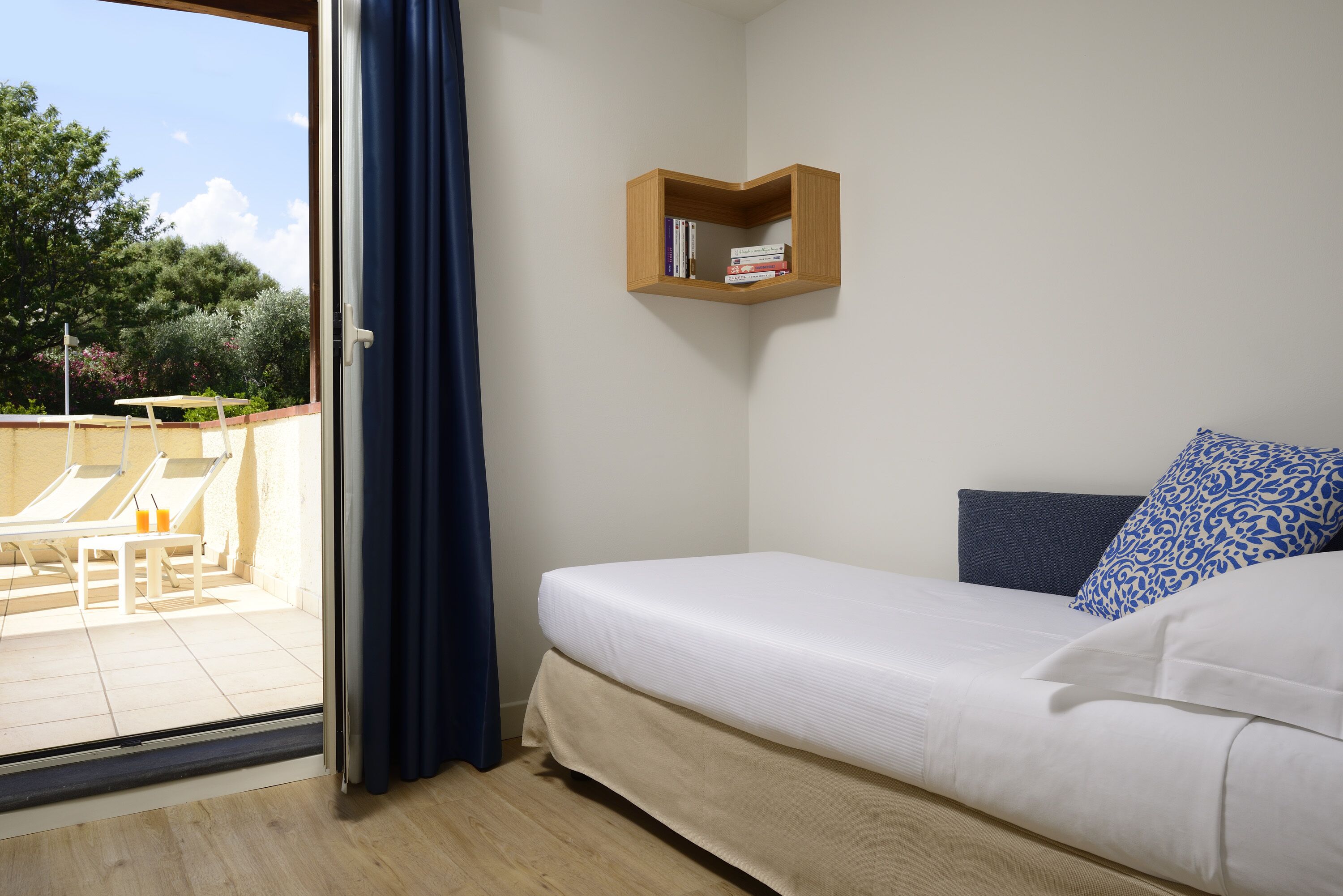 Ein Schlafzimmer mit Bett, einem kleinen Holzregal mit Büchern, einem Fenster mit Vorhängen und Blick auf eine Außenbereichs-Sitzgelegenheit.